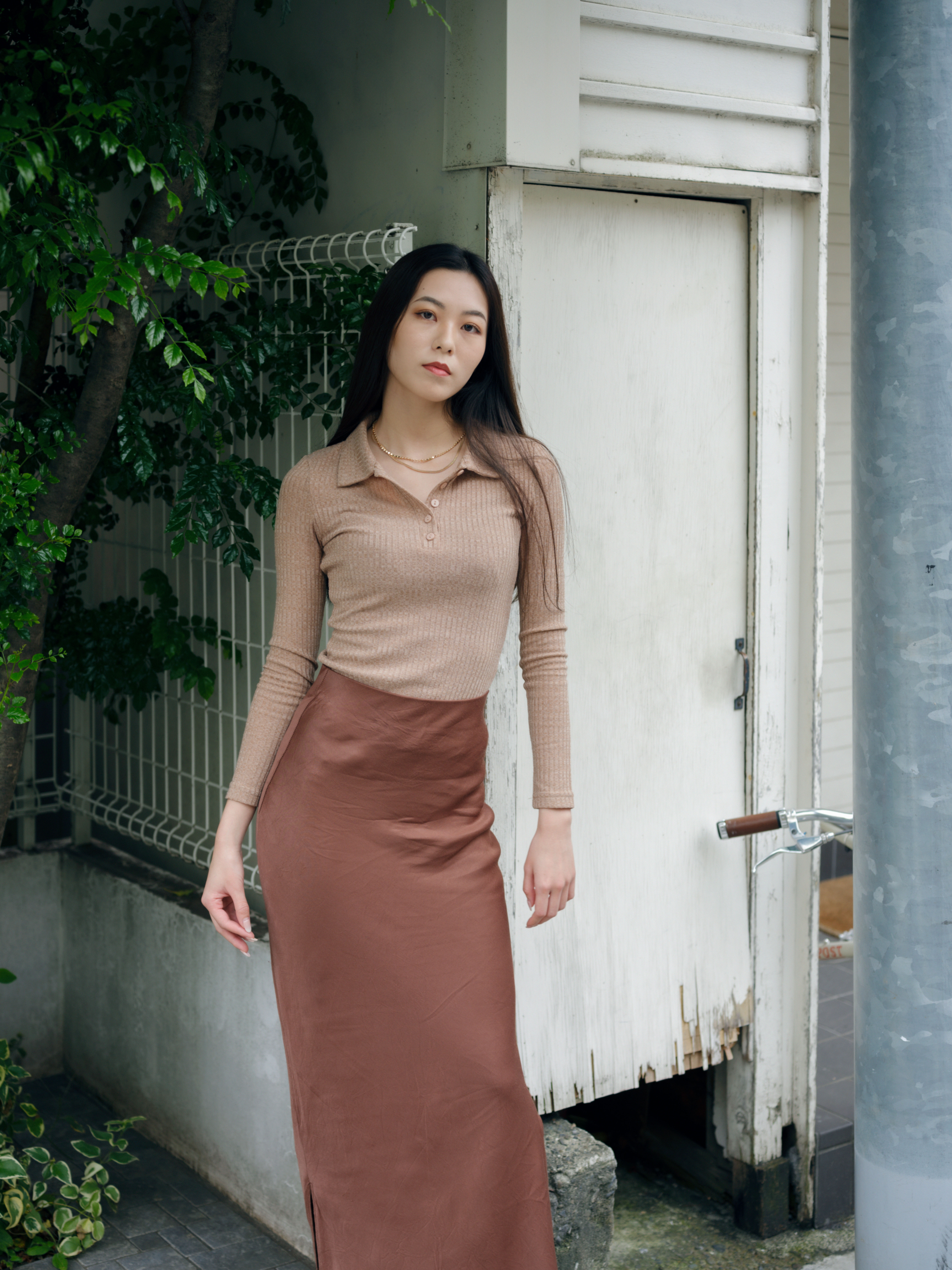 a woman with long straight hair wears a long brown skirt and a beige knit shirt with two gold chains.  she stands on a side street
