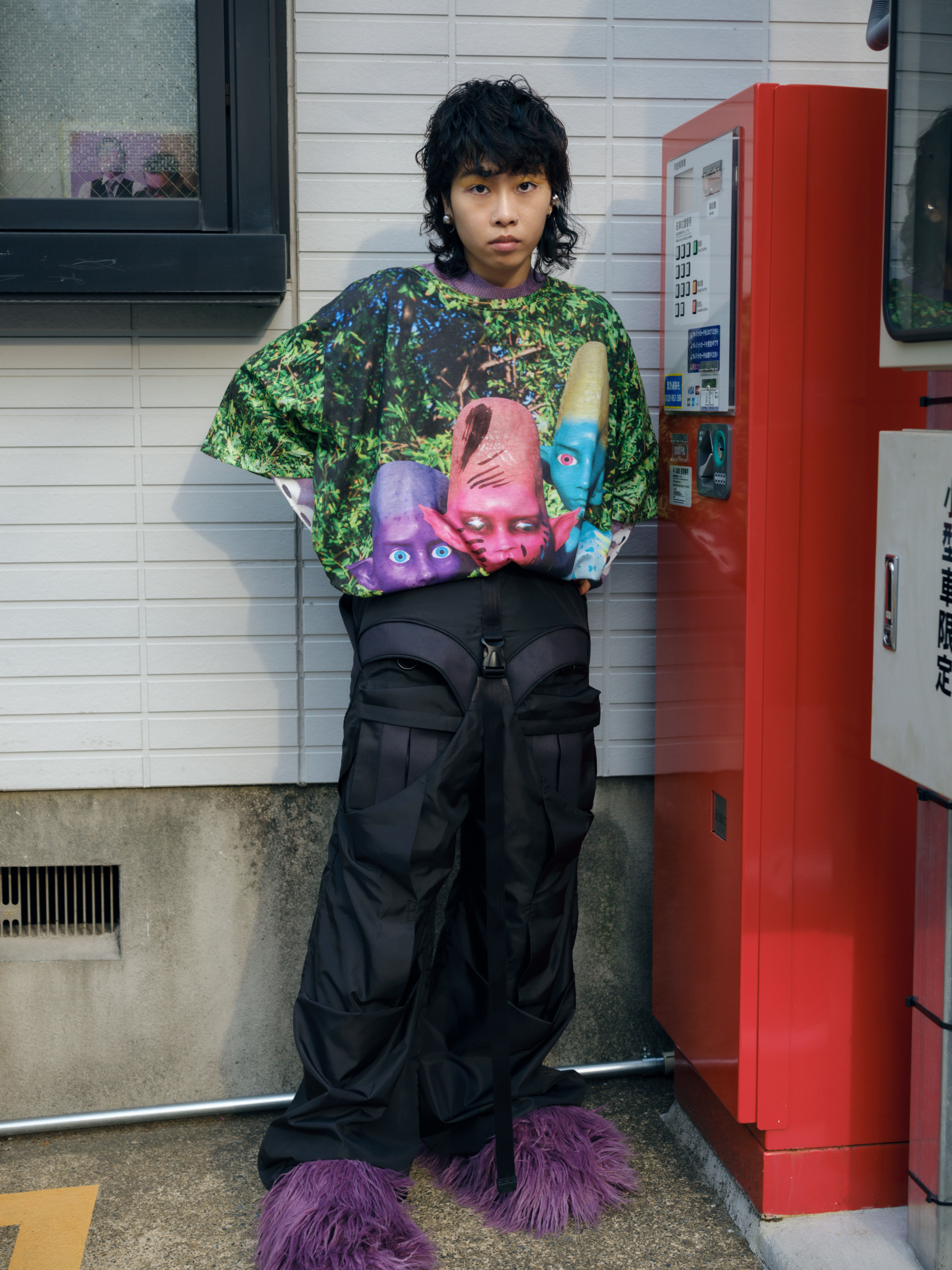 a person in a colourful print tshirt, combat trousers and big fluffy purple shoes stands by a vending machine