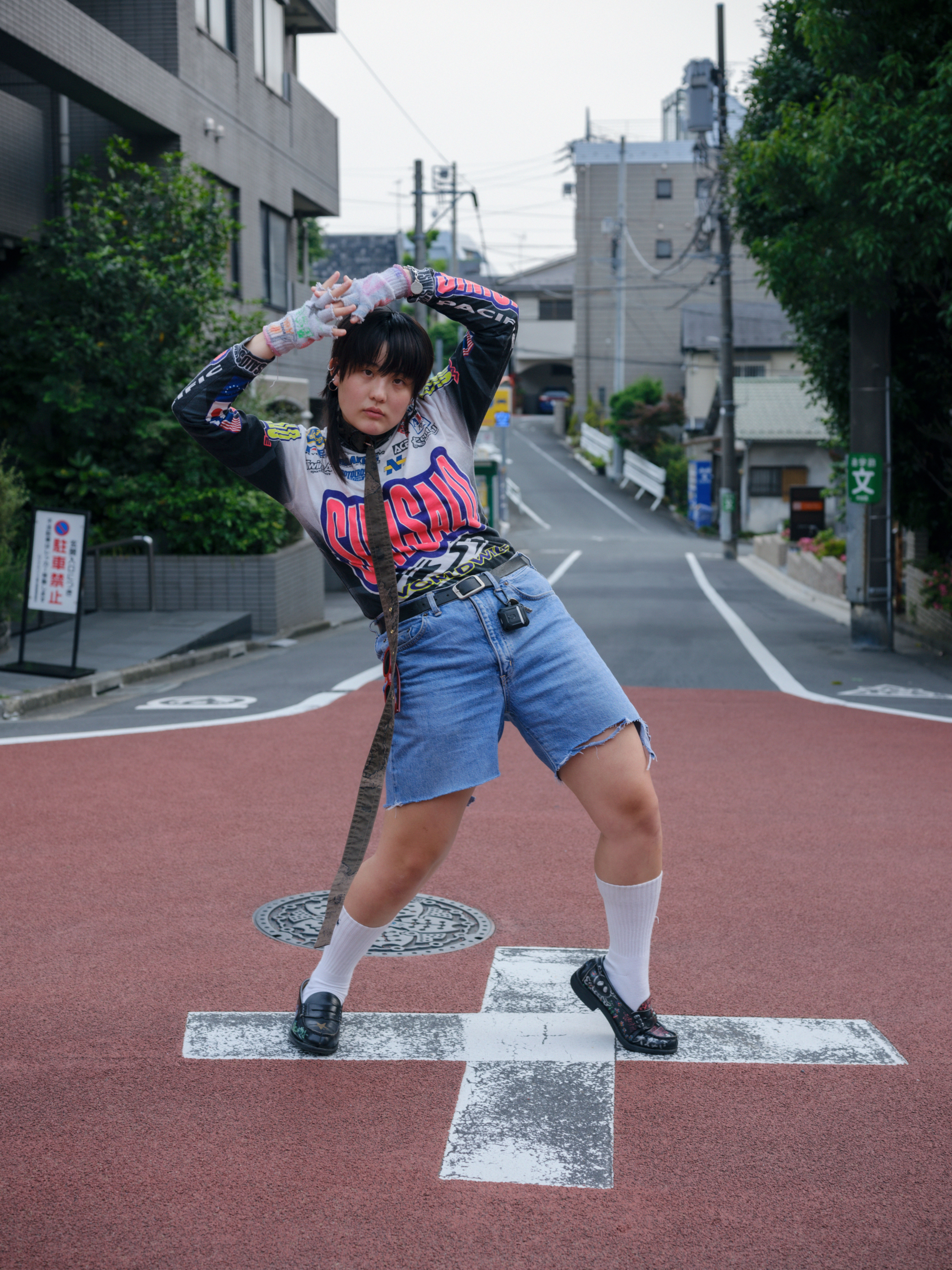 a person in a motocross shirt and denim shorts is interestingly leaning with his arms above his head in the middle of a tokyo residential street 