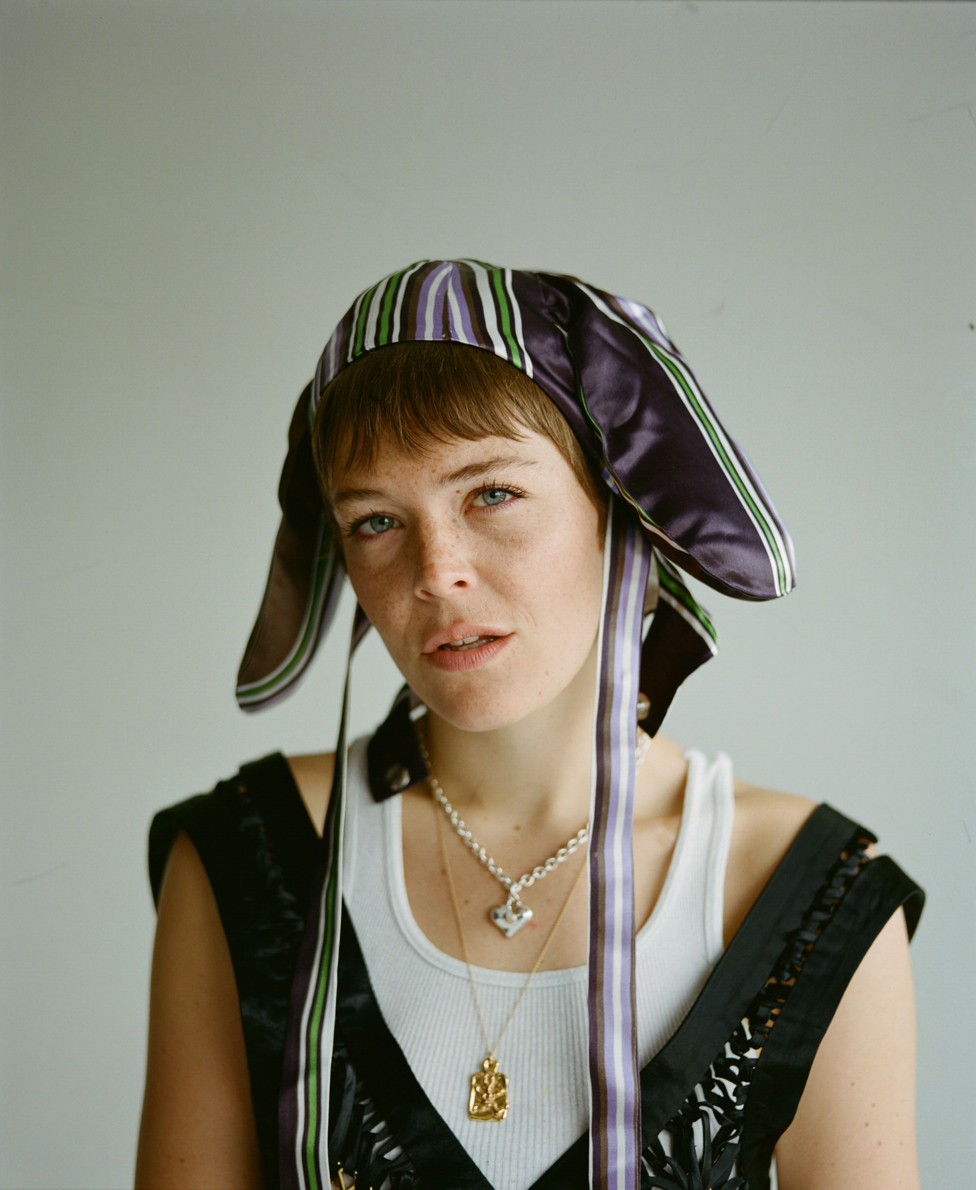 a facial close up of maggie rogers, wearing a silk trapper hat and a black vest over a white vest