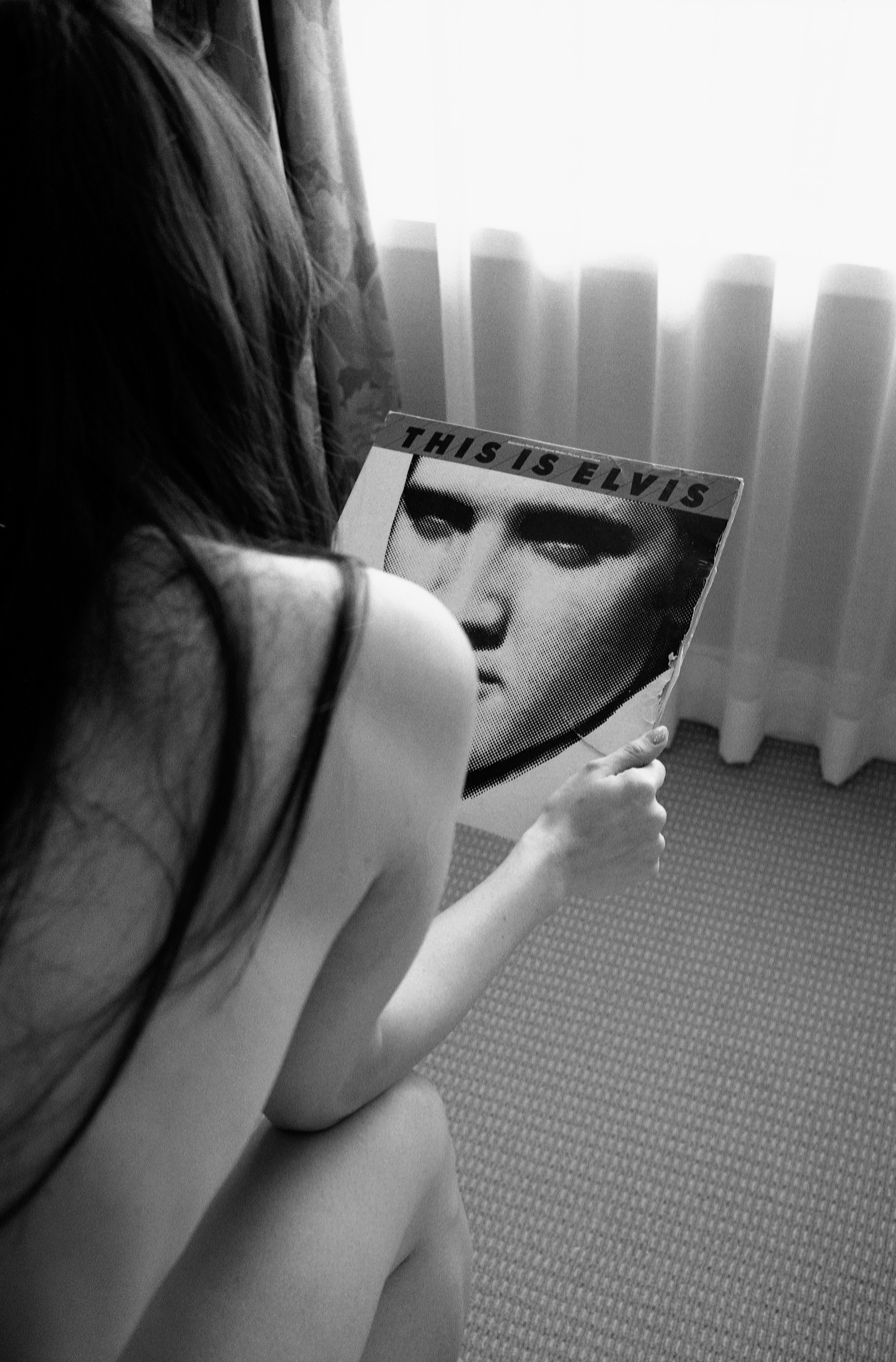 a long-haired nude woman holding an elvis vinyl sits on the edge of a bed, shot from behind 