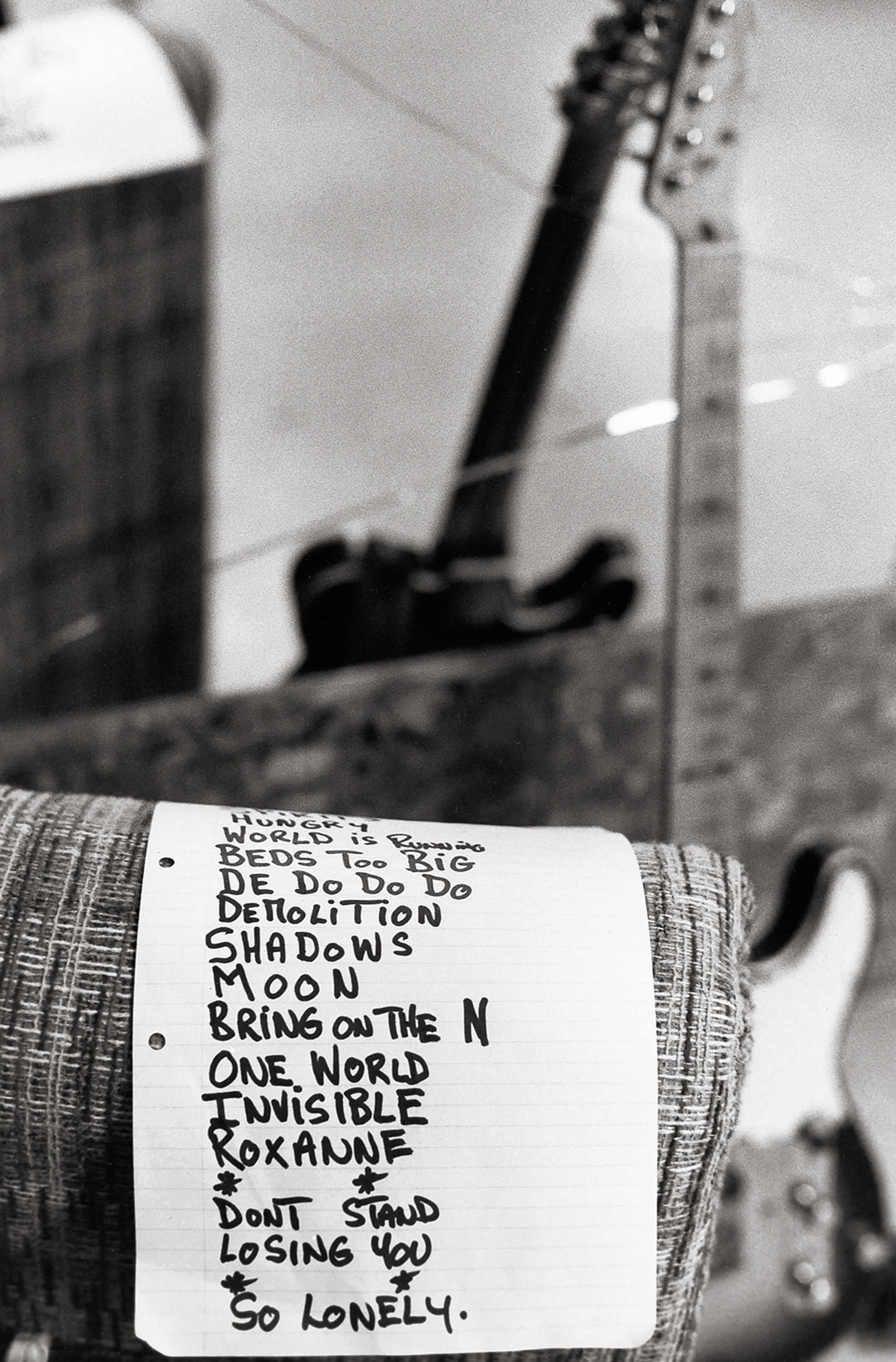 a handwritten set list rests over a cushion in a backstage room, a guitar stands out of focus in the background 