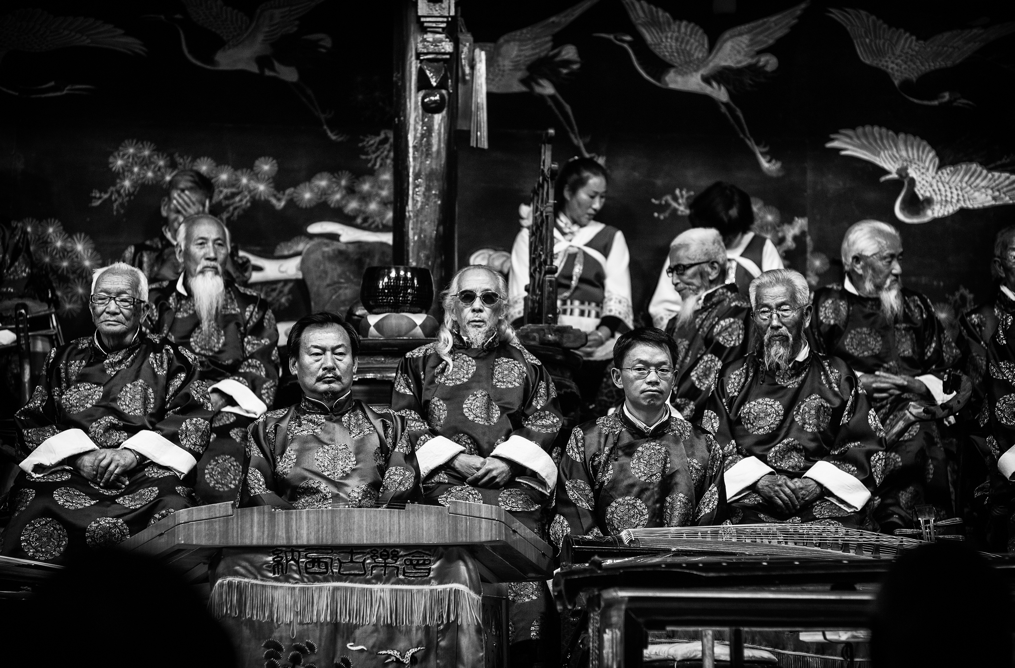 a group of elderly chinese men sit in a section of what appears to be a theatre in traditional dress