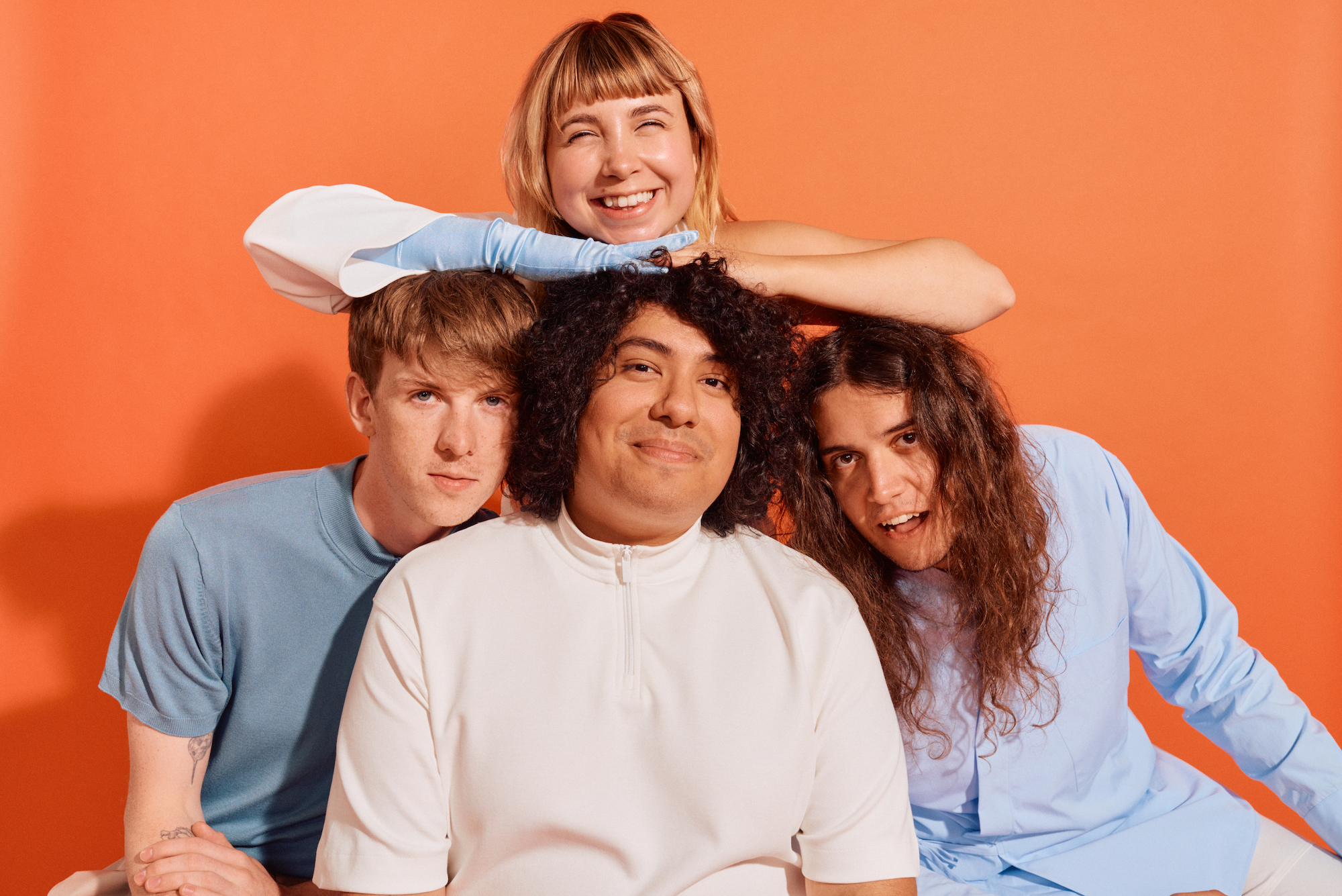 beach bunny band posing in white and blue shirts in front of an orange background