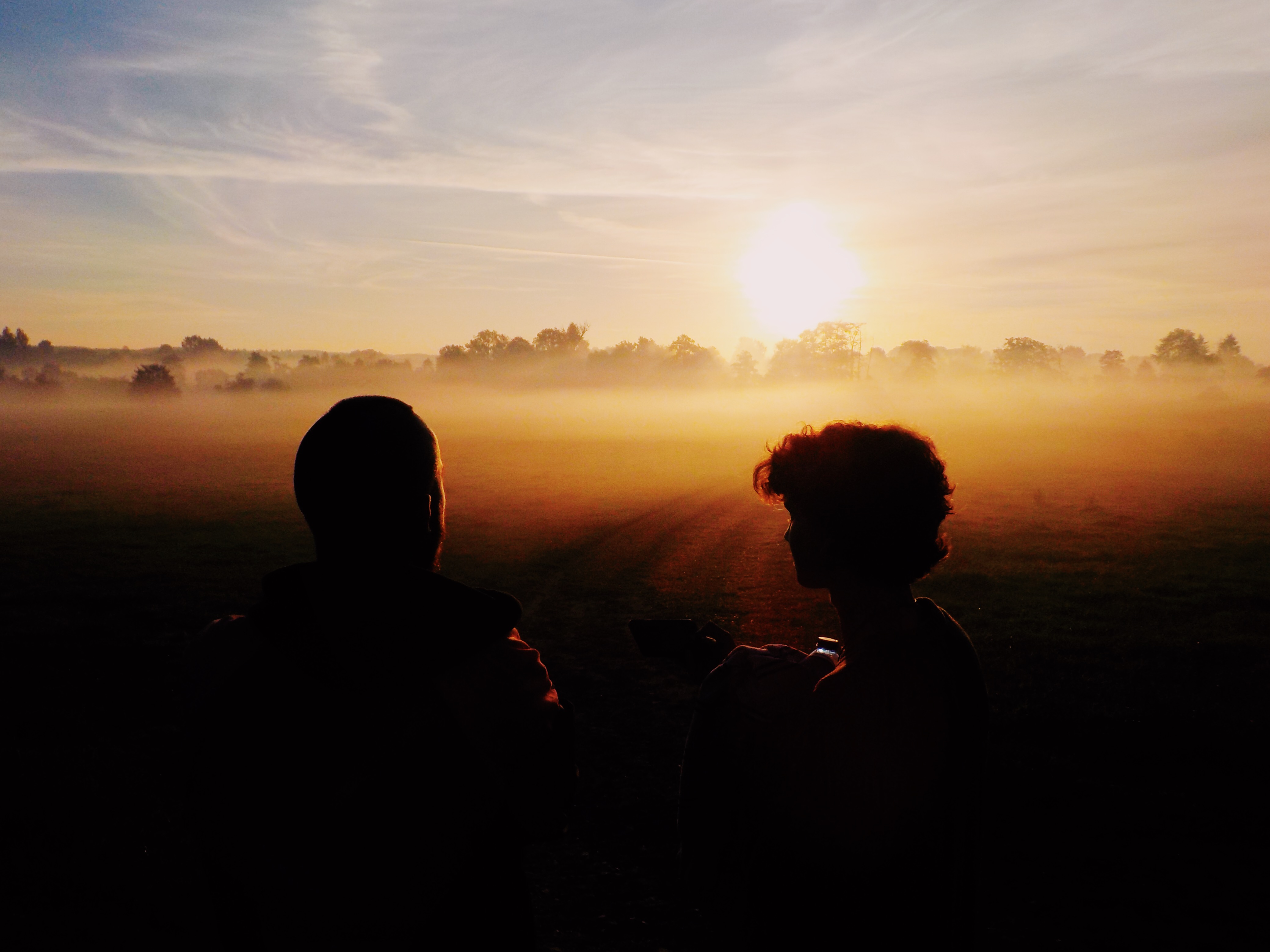 Two boys sitting on a field as the sun rises over the trees.