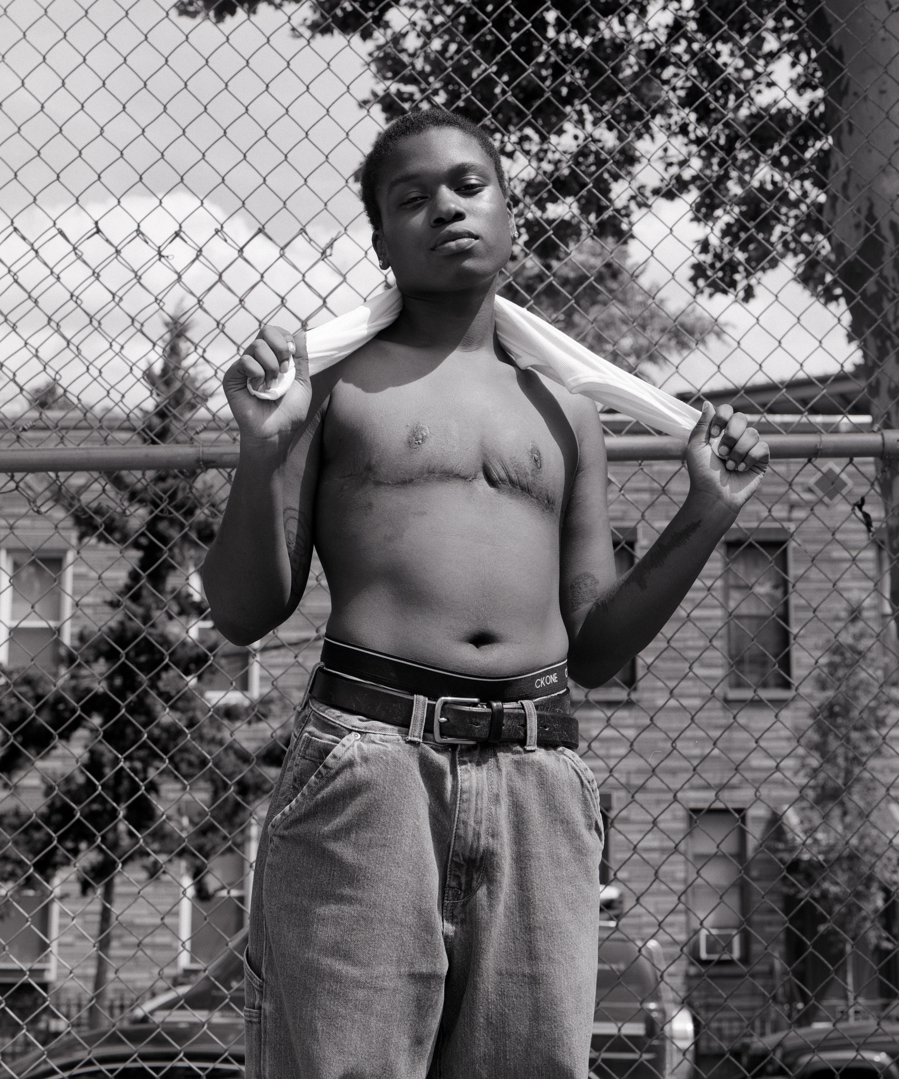 Person who has had top surgery standing outside a metal fence, shirtless in jeans, with a t-shirt held behind their neck
