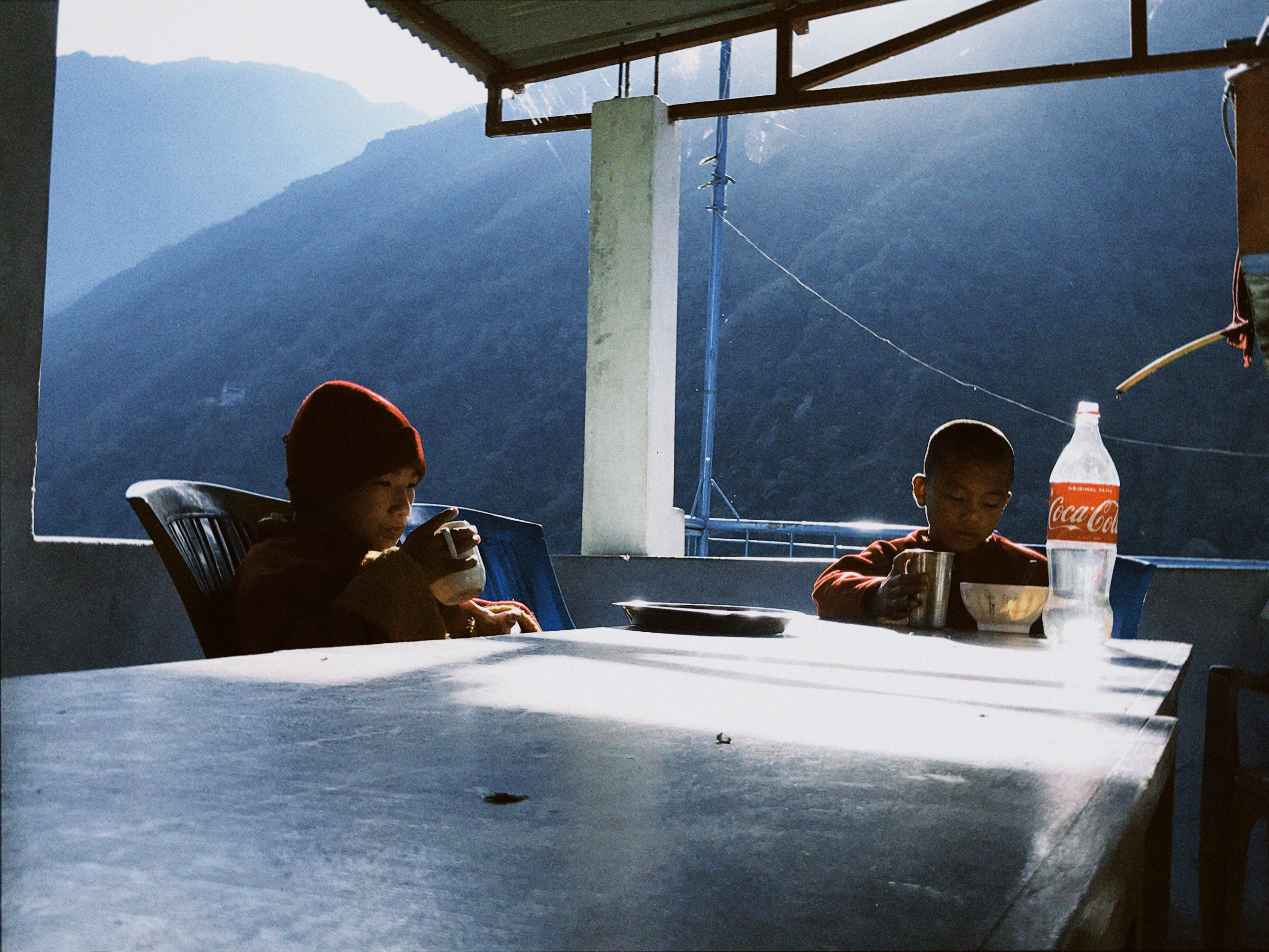 Two children drinking tea, having breakfast and water in a Coca Cola bottle at a table in a Tibetan monastery in the mountains