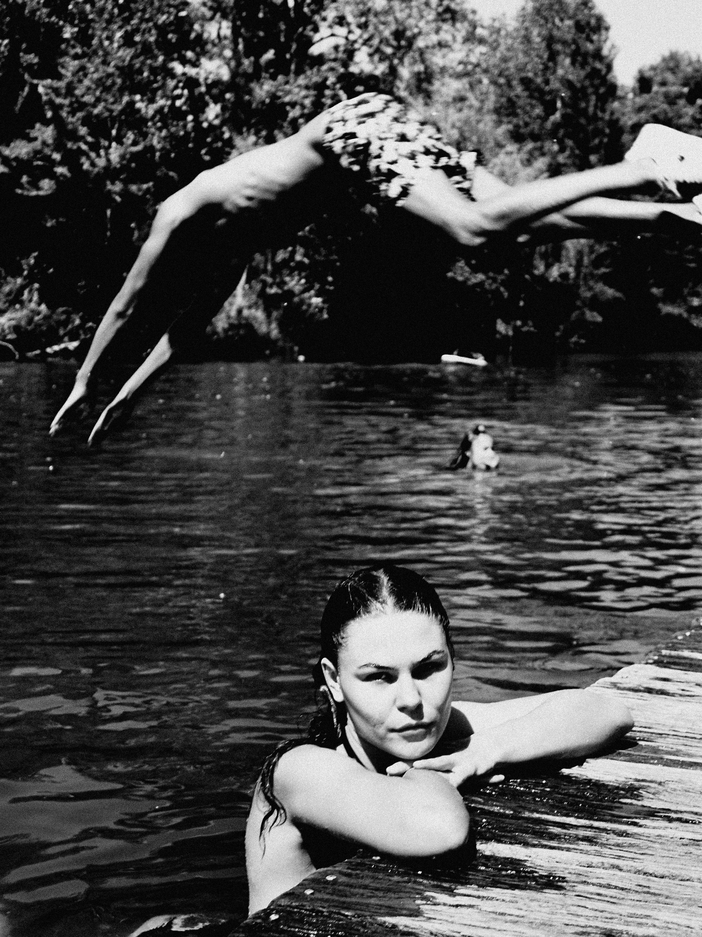 Girl in a lake leaning against the pier as someone else dives in the water
