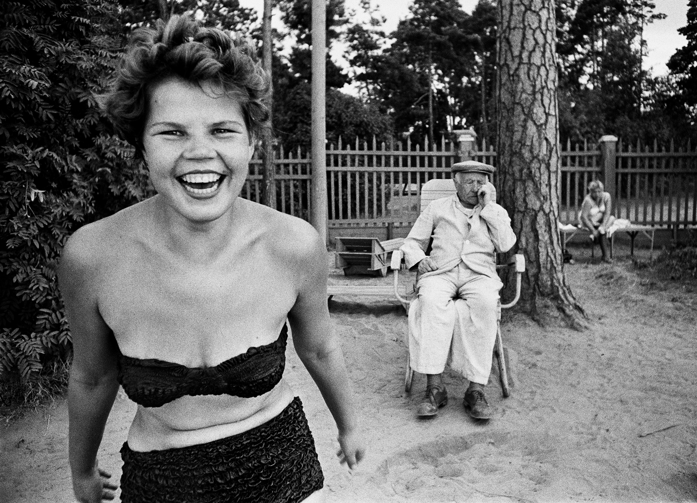 Black-and-white photo of a woman in a bikini outside laughing at the camera whilst an old man sleeps behind her