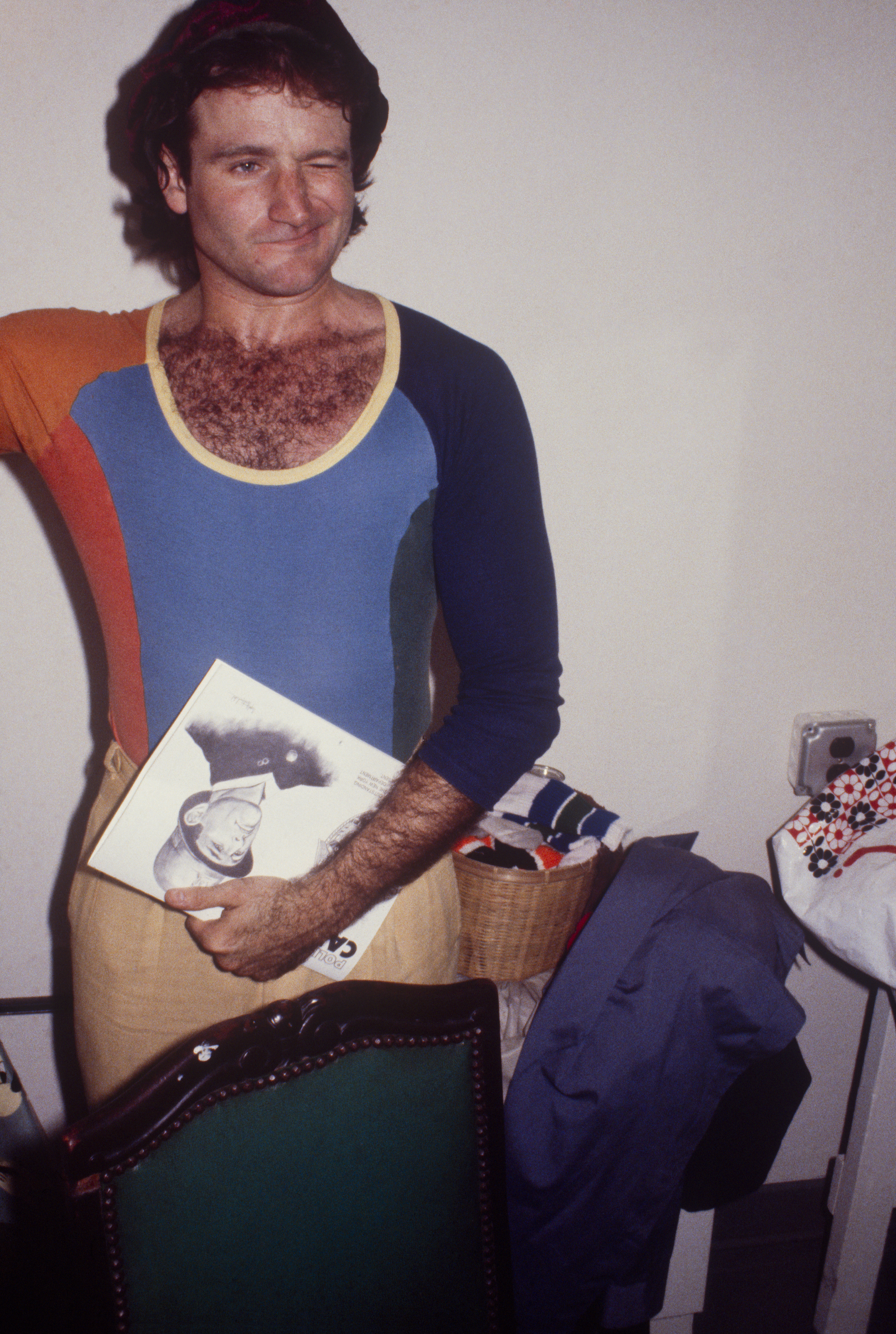 Robin Williams in a tight fitting multi-coloured top and beige trousers in a house holding a magazine and winking