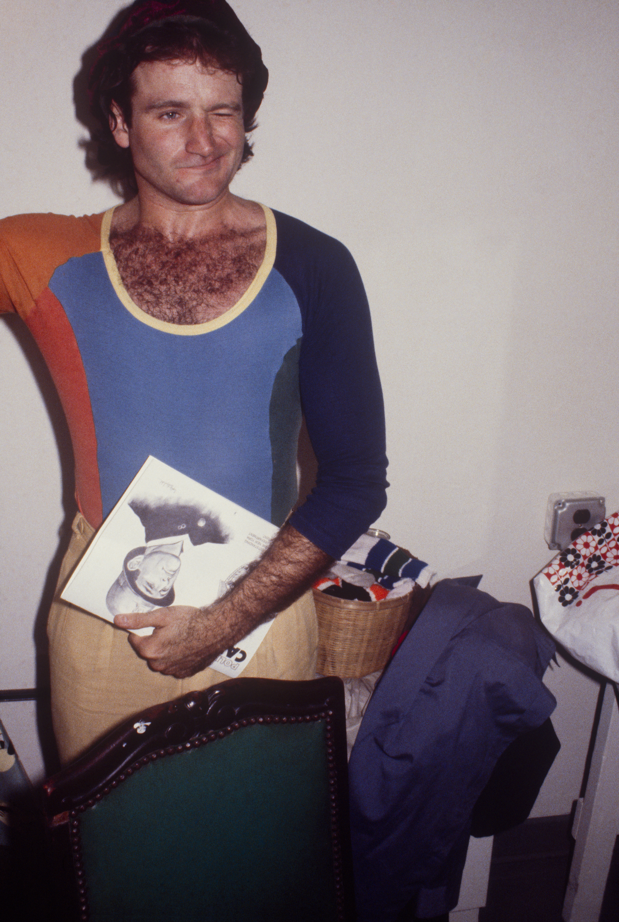 Robin Williams in a tight fitting multi-colored top and beige trousers in a house holding a magazine and winking