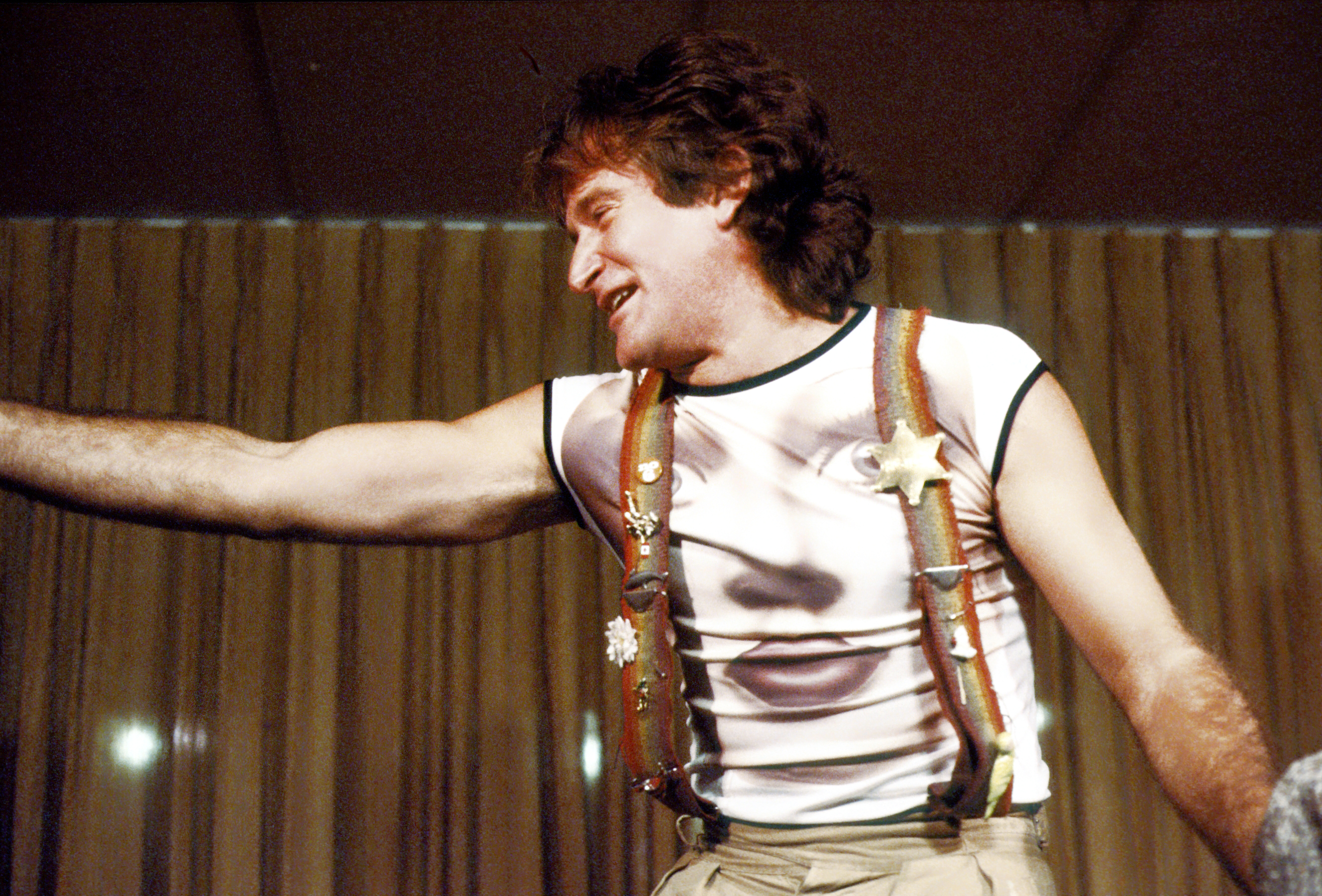 Robin Williams in rainbow suspenders and a face printed t-shirt performing on stage at the Roxy Theatre in 1979