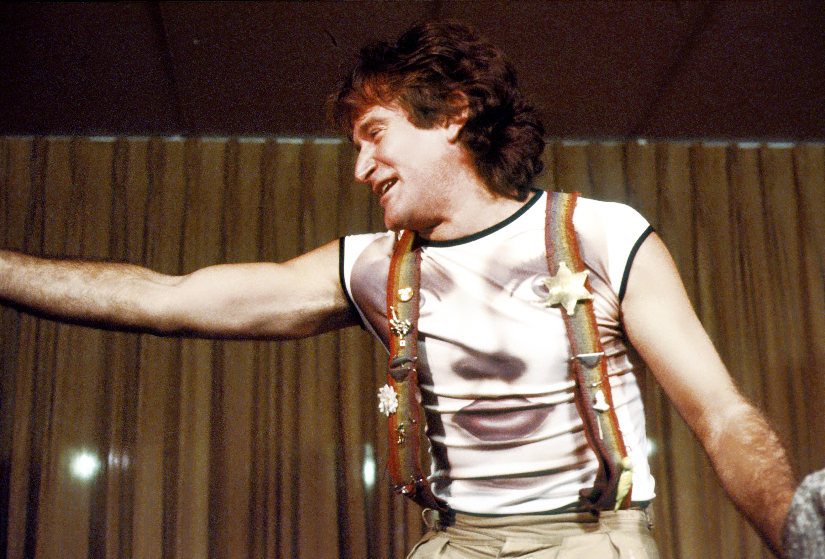 Robin Williams in rainbow suspenders and a face printed t-shirt performing on stage at the Roxy Theater in 1979