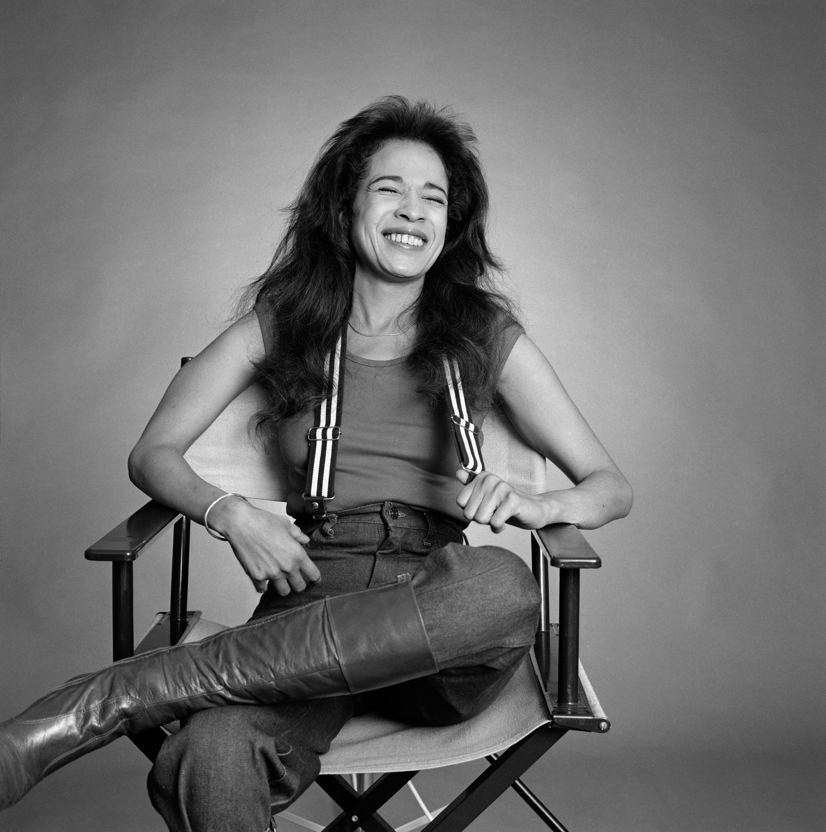 ronnnie spector sitting in a director's chair laughing wear suspenders, jeans and leather boots 1977