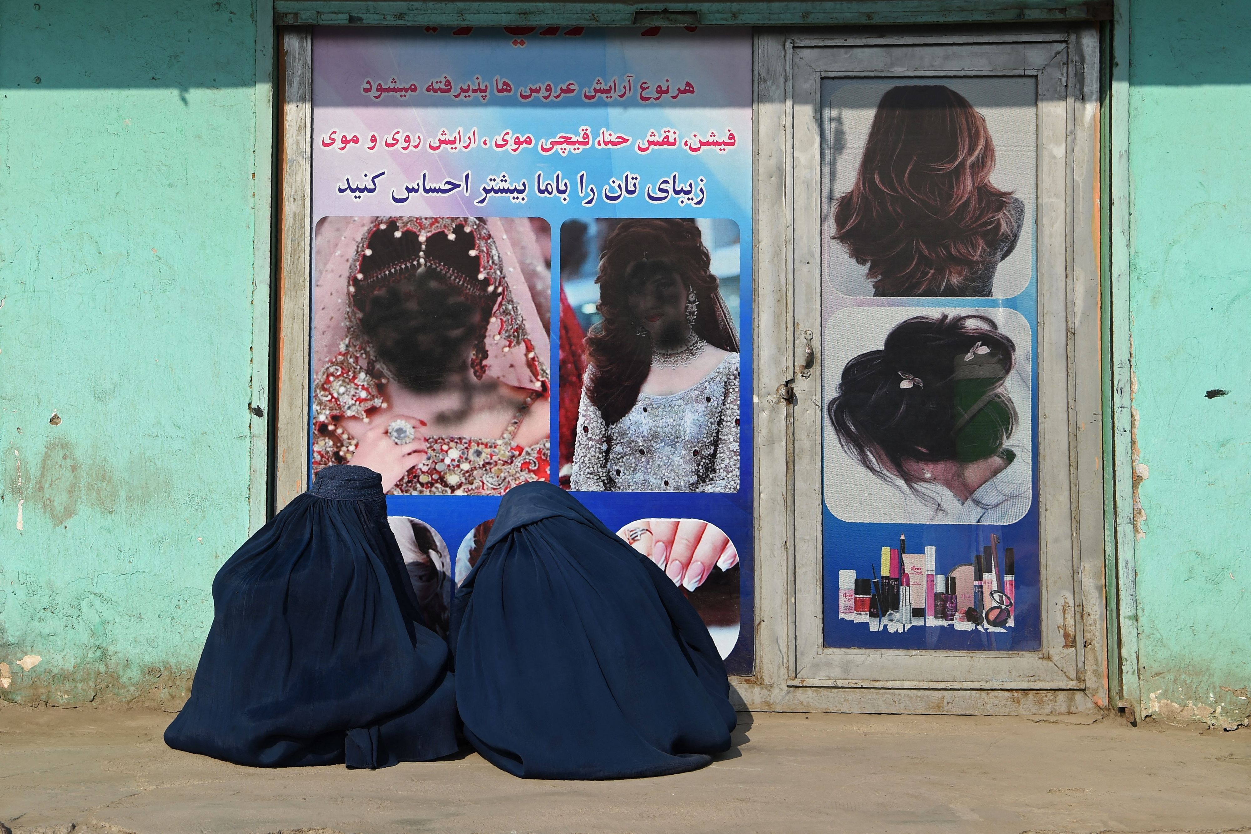 Dua perempuan dalam balutan burkak duduk di depan salon kecantikan di Jalalabad pada 13 Desember 2021. Poster perempuan telah ditimpa cat hitam. Foto: Wakil Kohsar / AFP