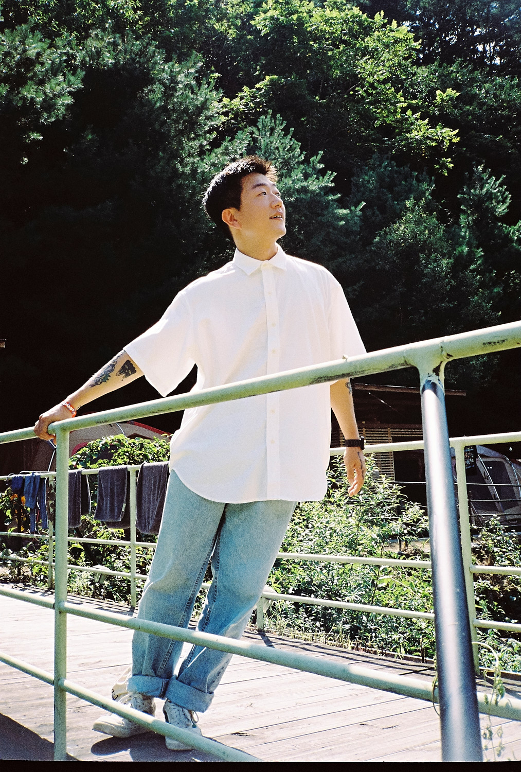 a young man wearing light jeans and a white shirt leans to one side on a bridge; trees are behind him