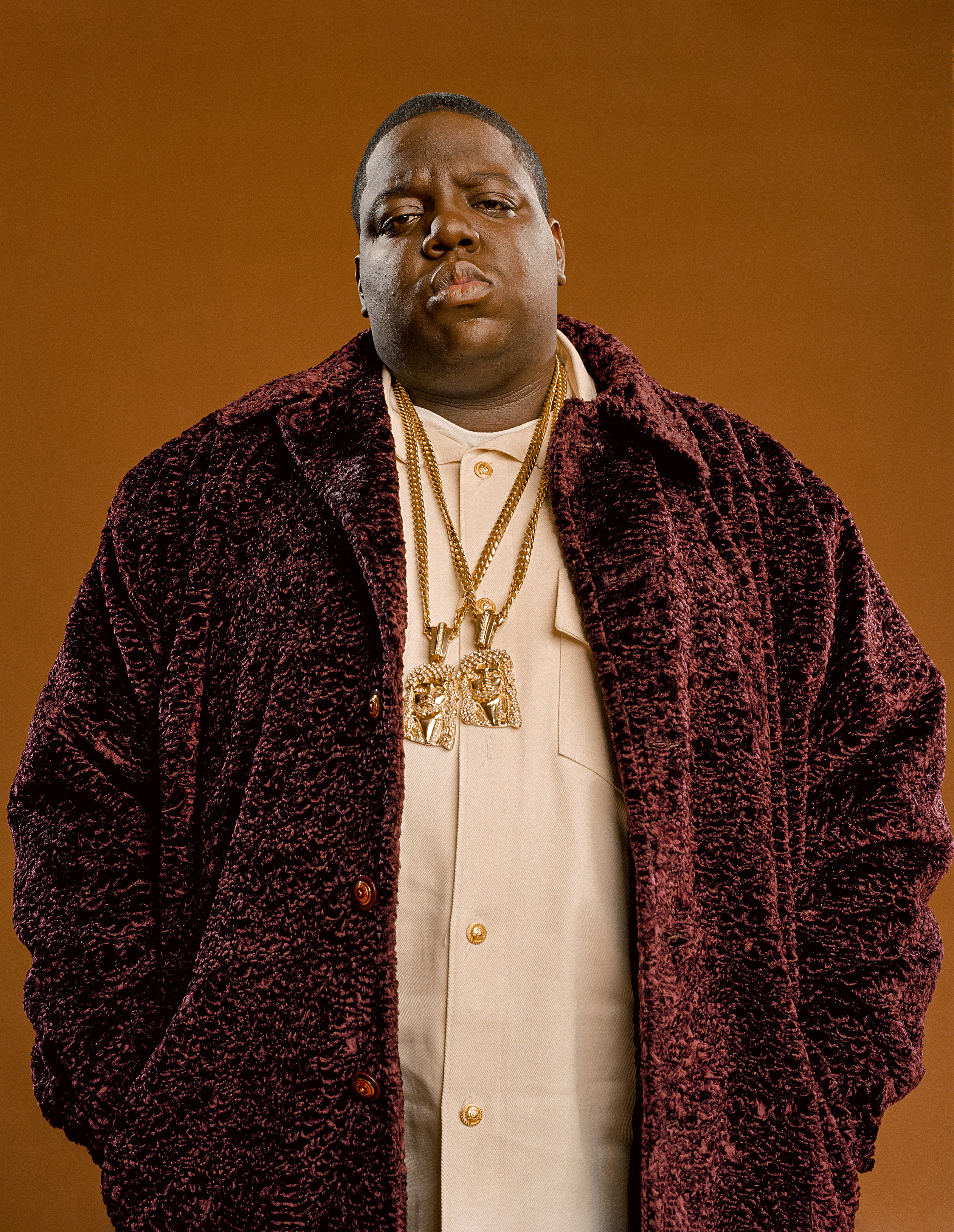 Der Rapper Notorious B.I.G. steht vor einem braunen Hintergrund. Er trägt zwei Jesus-Ketten und eine bordeauxfarbene Jacke. Er schaut in die Kamera