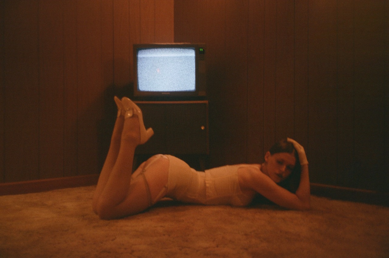 ethel reclines on a carpeted floor in white lingerie beneath an analog television set