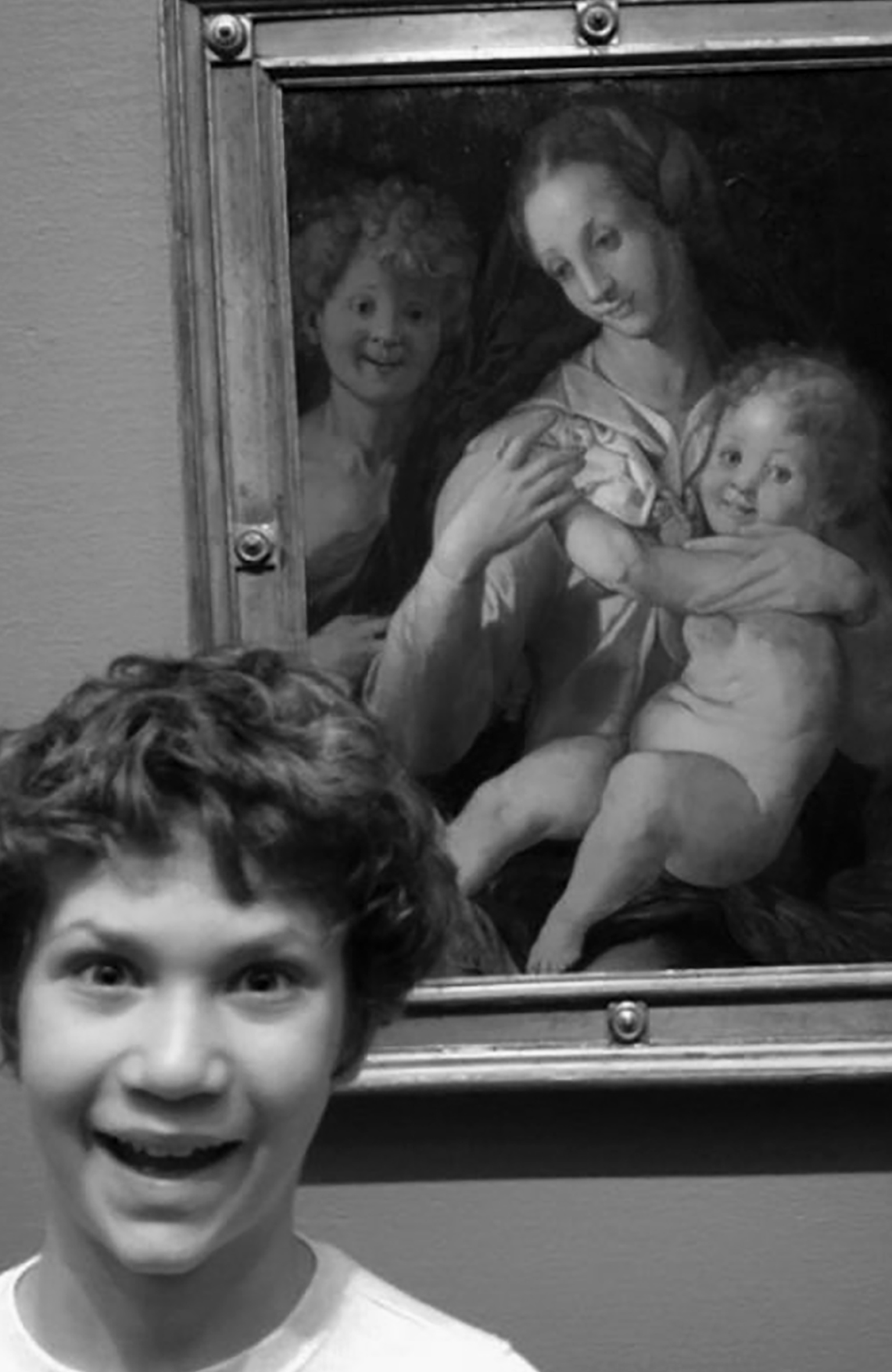 Bocah laki-laki berambut ikal memamerkan ekspresi konyol di depan lukisan anak laki-laki berambut ikal dengan ekspresi konyol