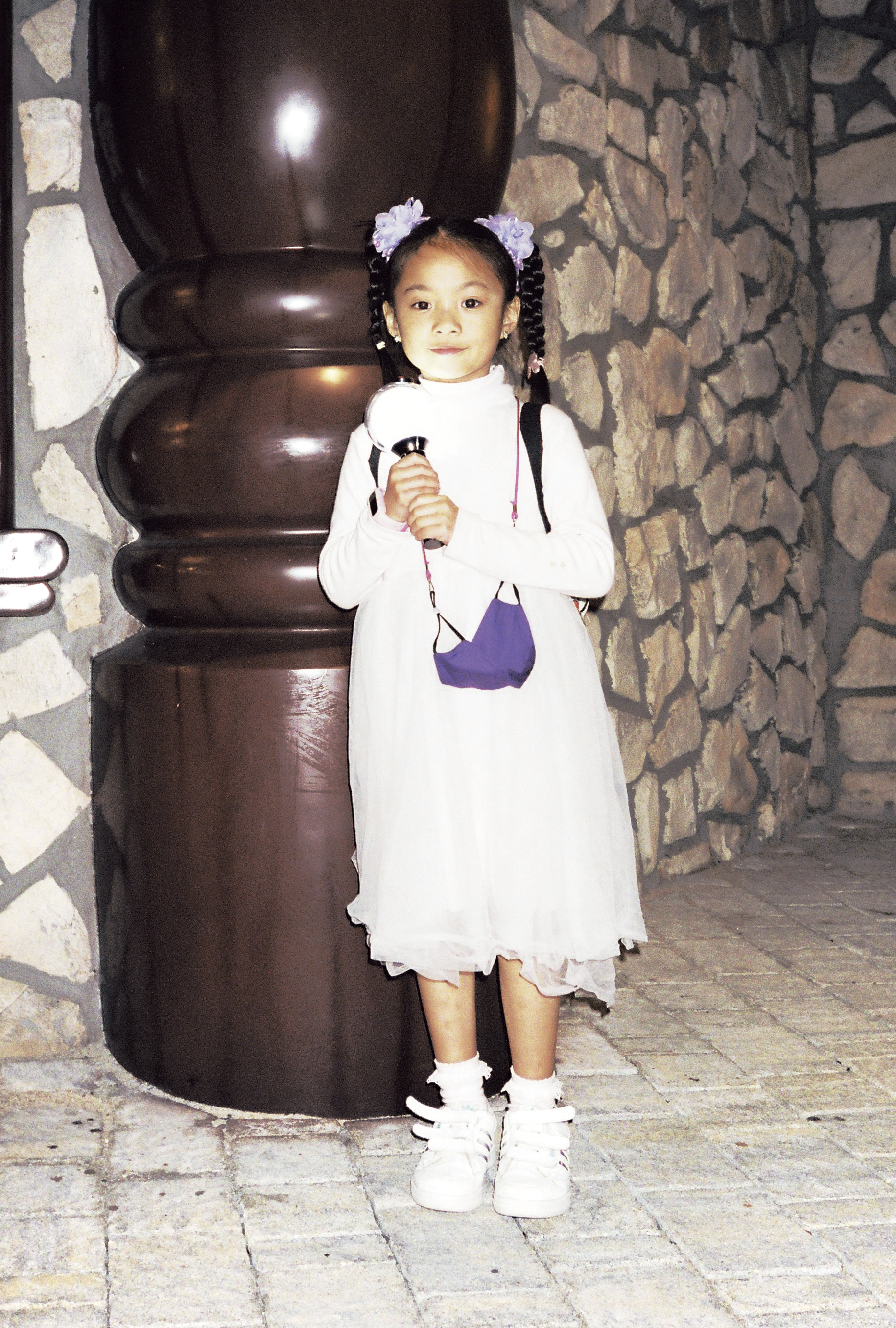 a young girl in a white dress and high pigtails clutches a light toy, a face mask hanging around her neck