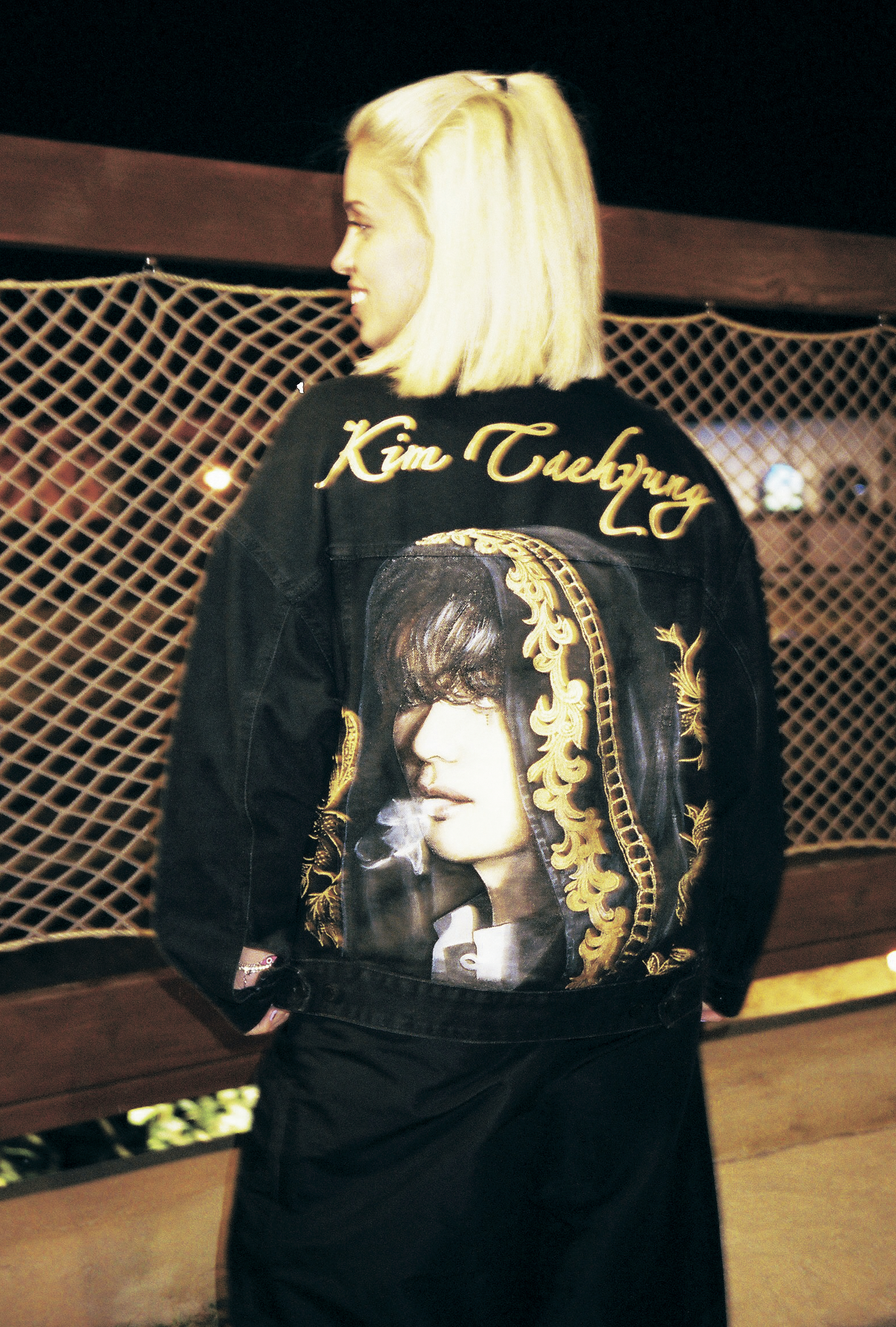 a blonde model turns to show the back of her jacket, featuring a gold embossed portrait of kim taehyung
