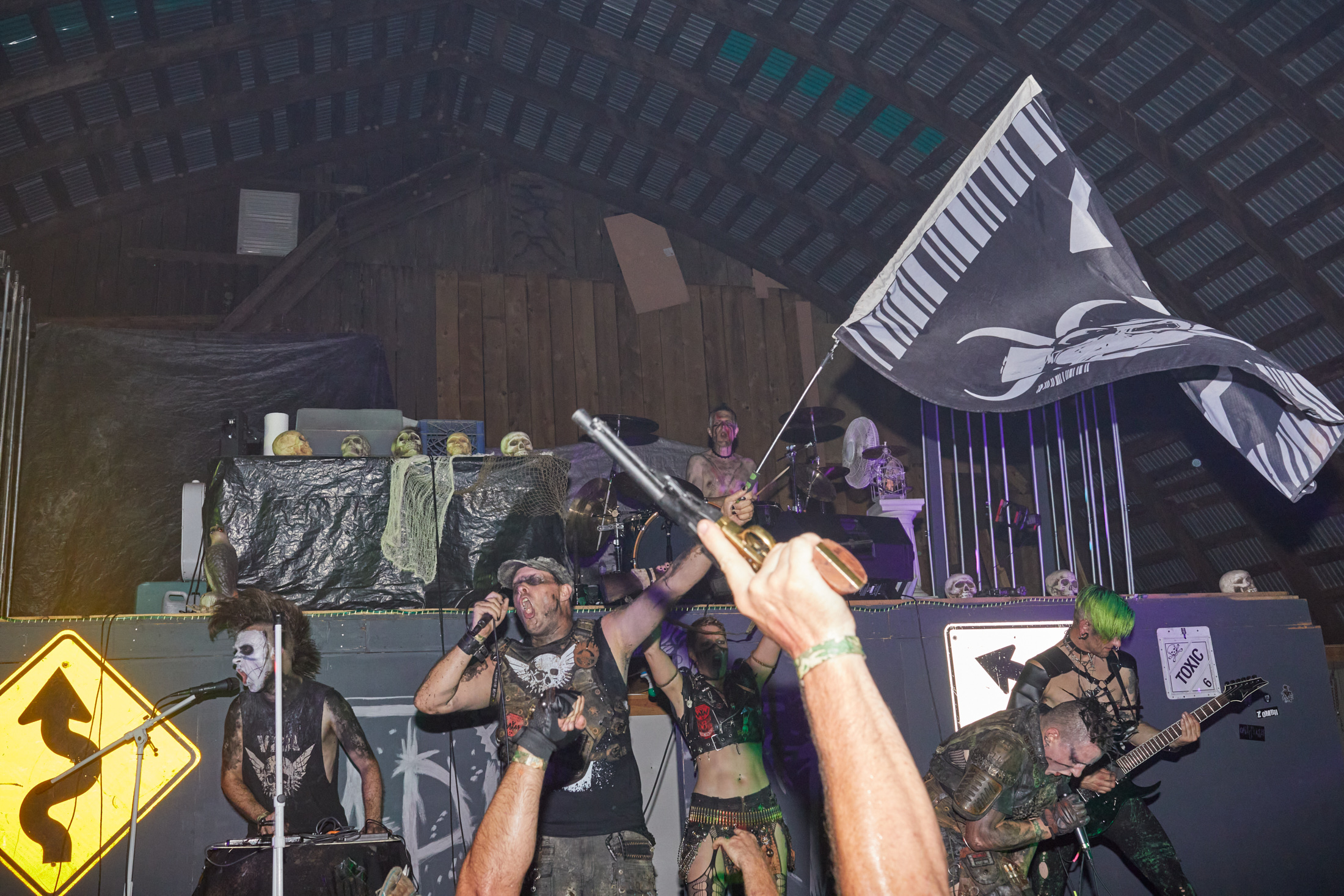 A band plays on a stage inside a barn, an attendee's prop gun is raised in the foreground