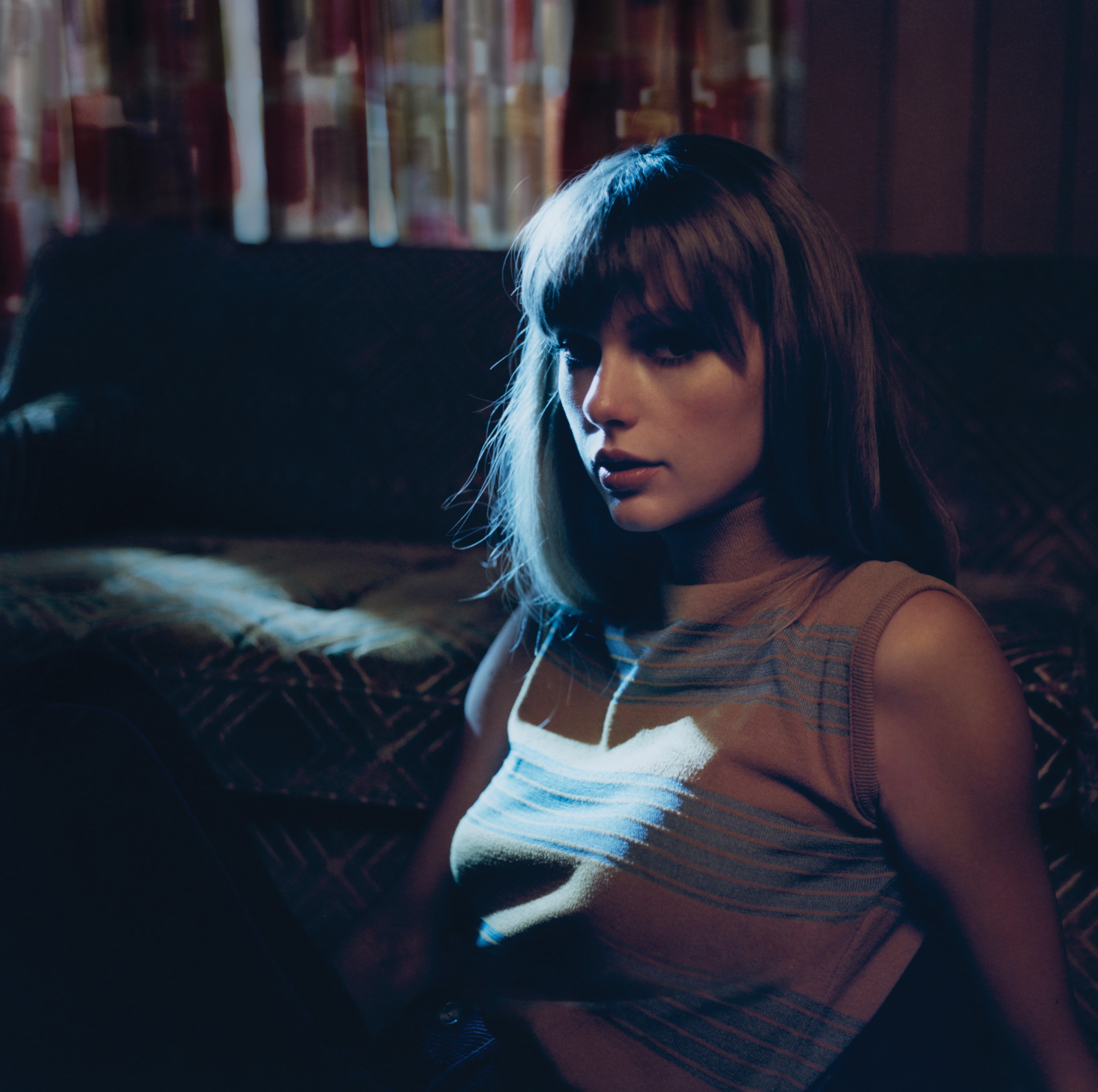 taylor sits in front of a couch in a dark room wearing a striped tank top
