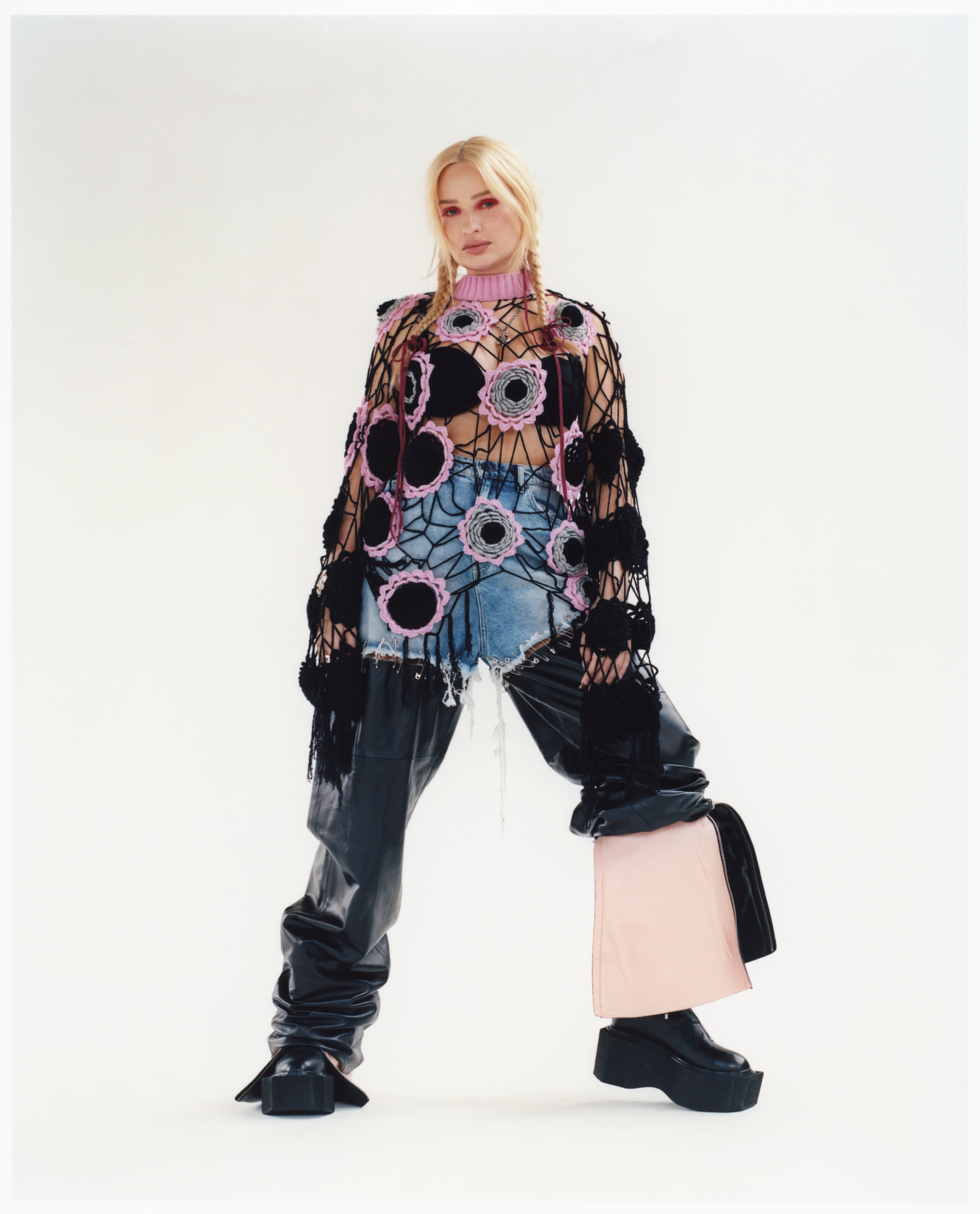 full length portrait of kim petras wearing pink eyeshadow and her hair in plaits and an outfit comprised of a knitted holy sweater and leather trousers 