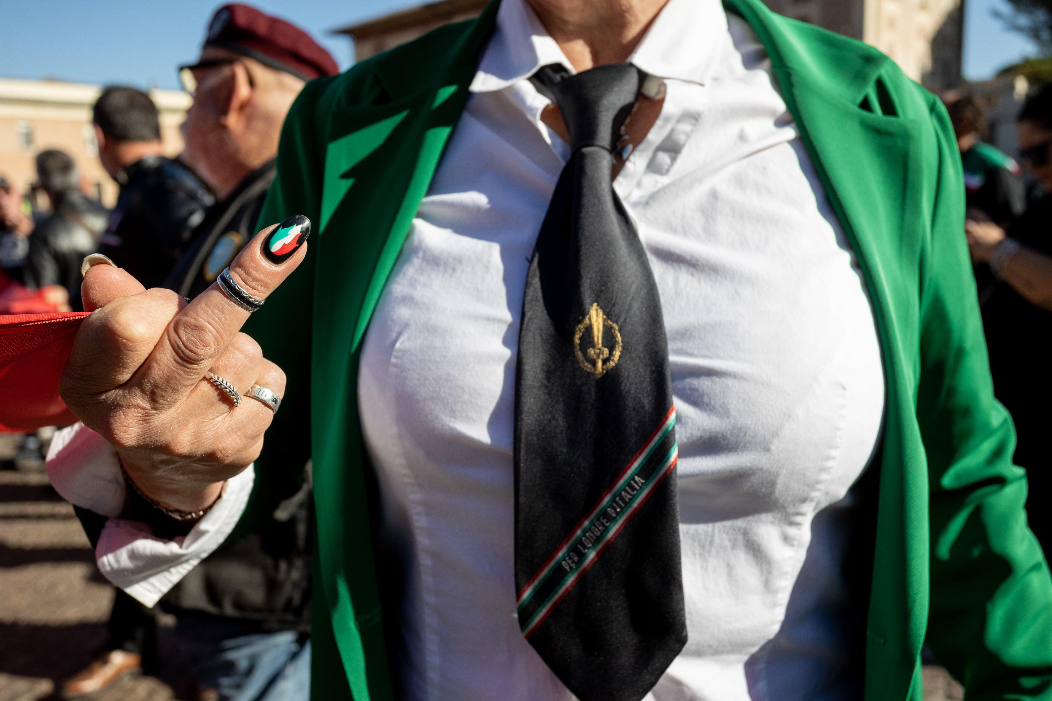 Perempuan mengenakan pakaian dengan warna bendera Italia. Dasi yang ia kenakan bertuliskan slogan fasis “Per L’Onore D’Italia” (“Demi Kehormatan Italia”).