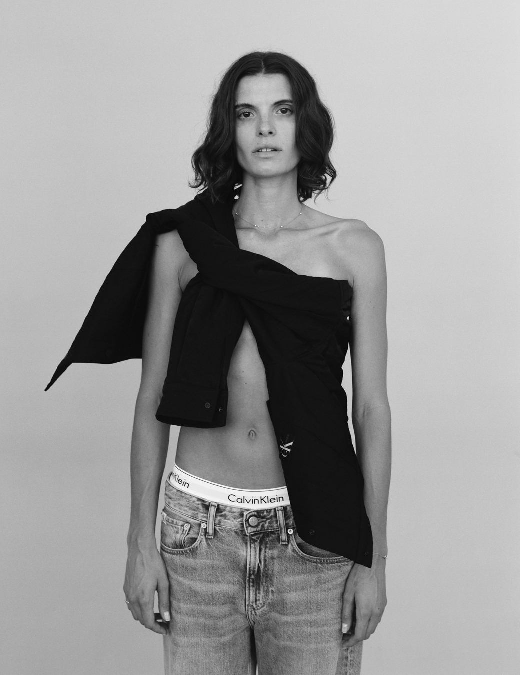 a model wears a black shirt tied across her chest with a pair of jeans, the waistband of her CK underwear showing