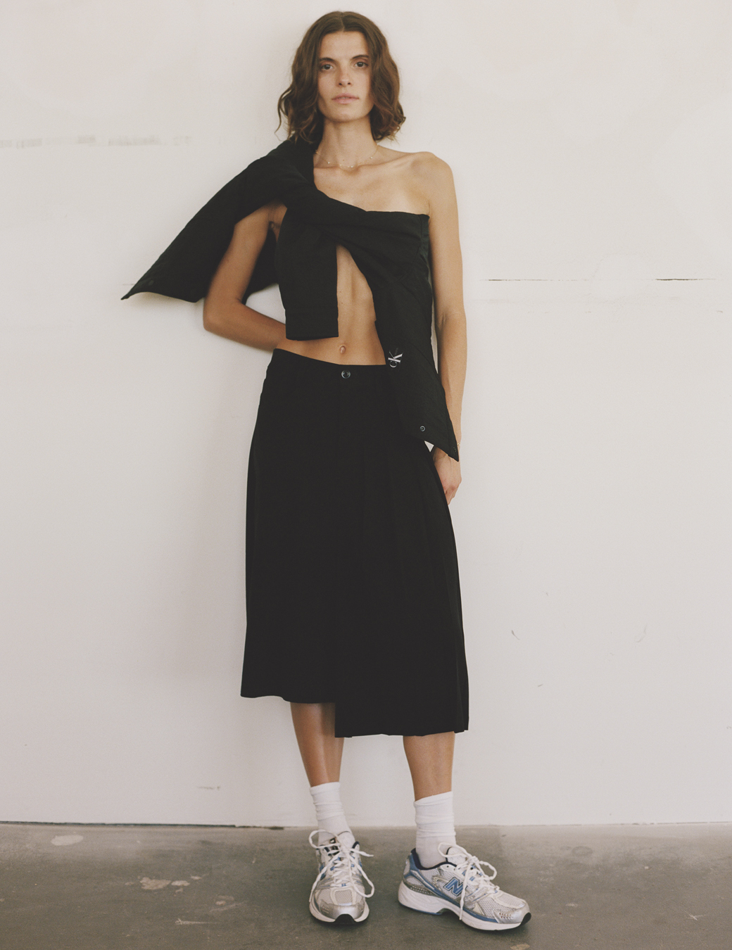 a model wears a black midi skirt with a shirt tied across her chest and a pair of old school sneakers