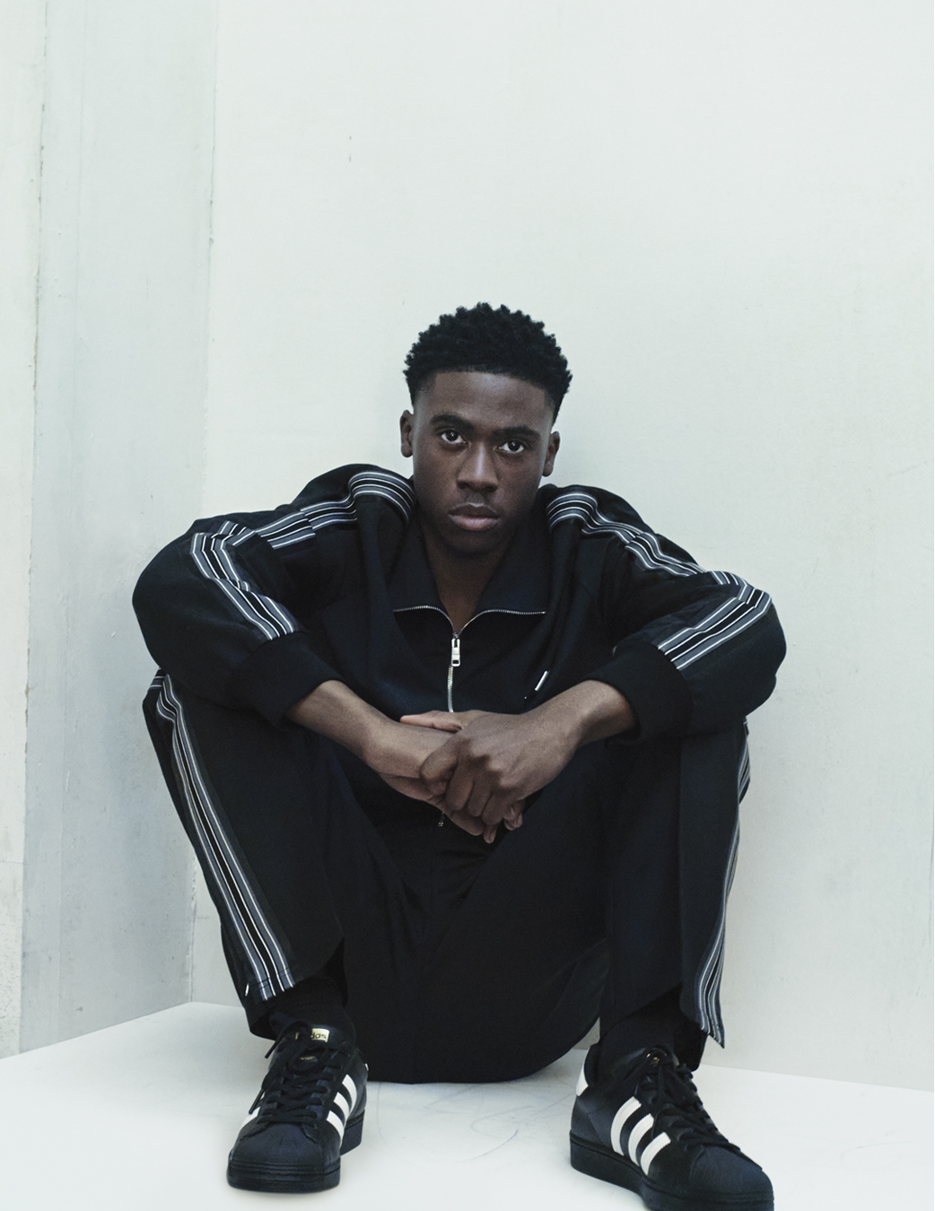 Nino in a tracksuit sat in a concrete white room photographed by Dale Cutts for i-D’s The Royalty Issue, no. 370, Winter 2022