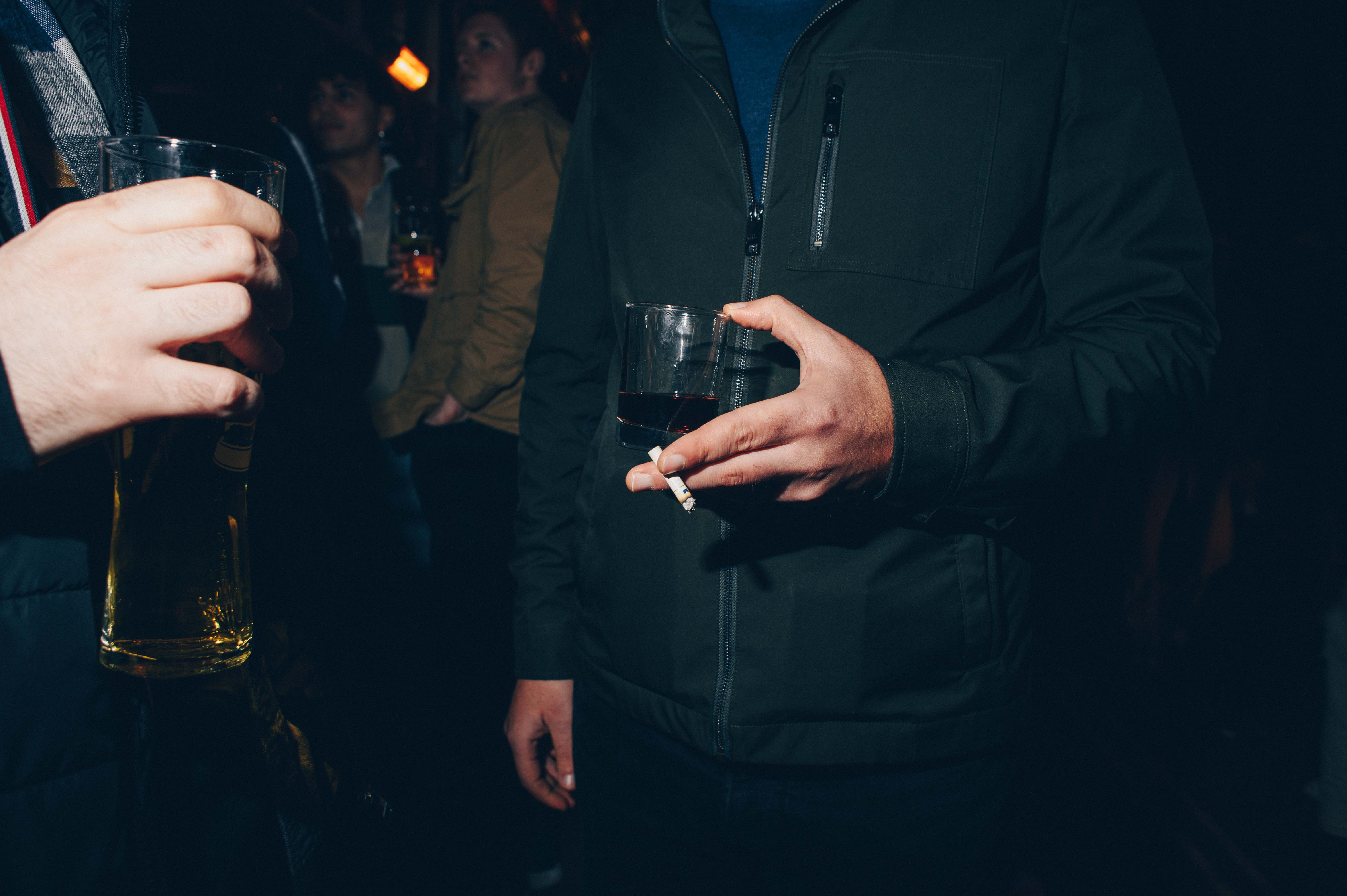 A detail shot of a man holding a small glass of alcohol with a cigarette in-between his fingers.