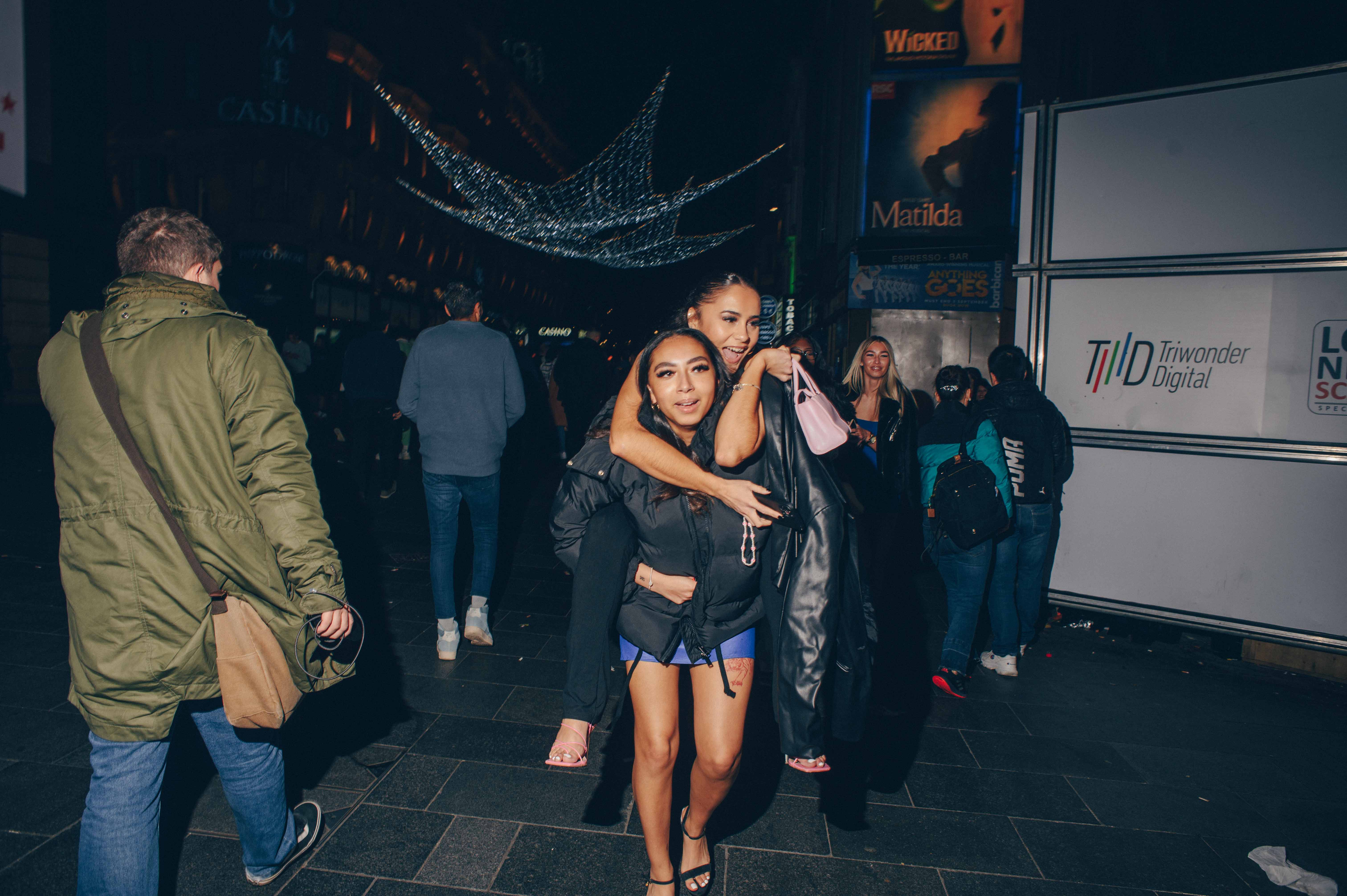 A woman gives another woman a piggyback ride on a busy street.