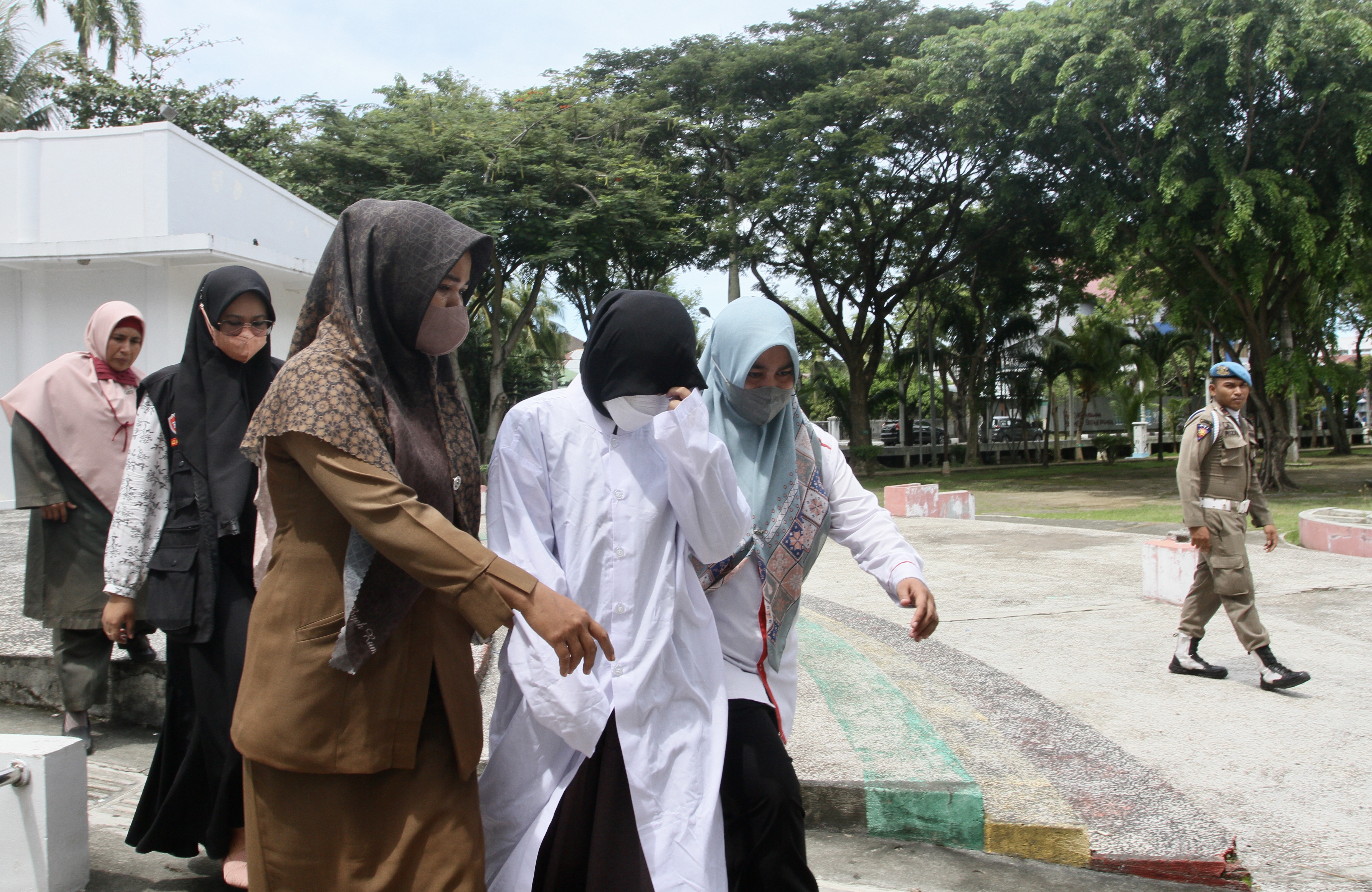 Perempuan bergamis putih yang mengenakan masker putih dan jilbab hitam dituntun dua orang perempuan