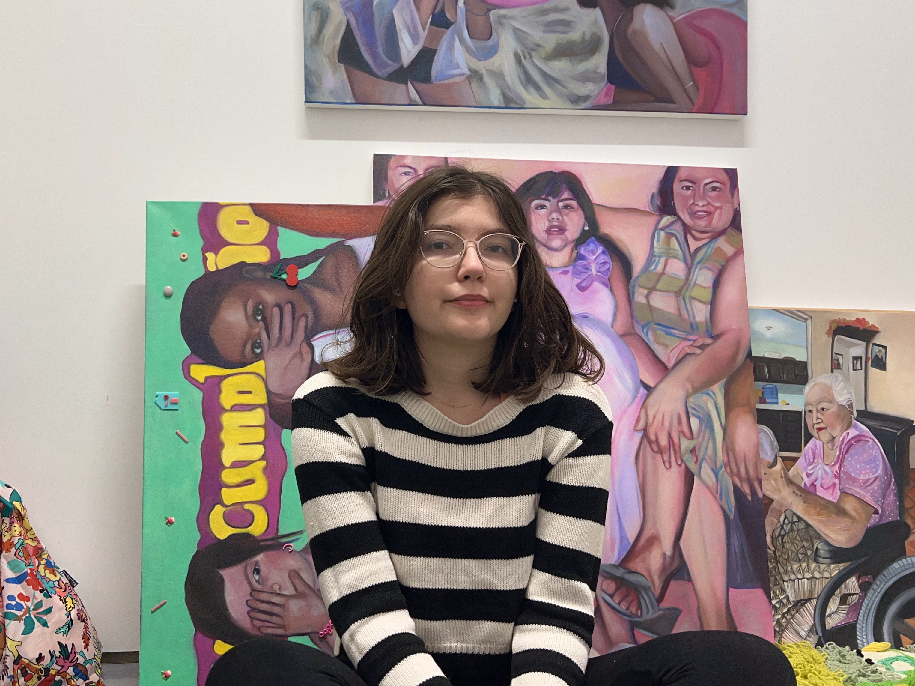 A young woman in a white and black striped shirt sits in front of painted canvasses