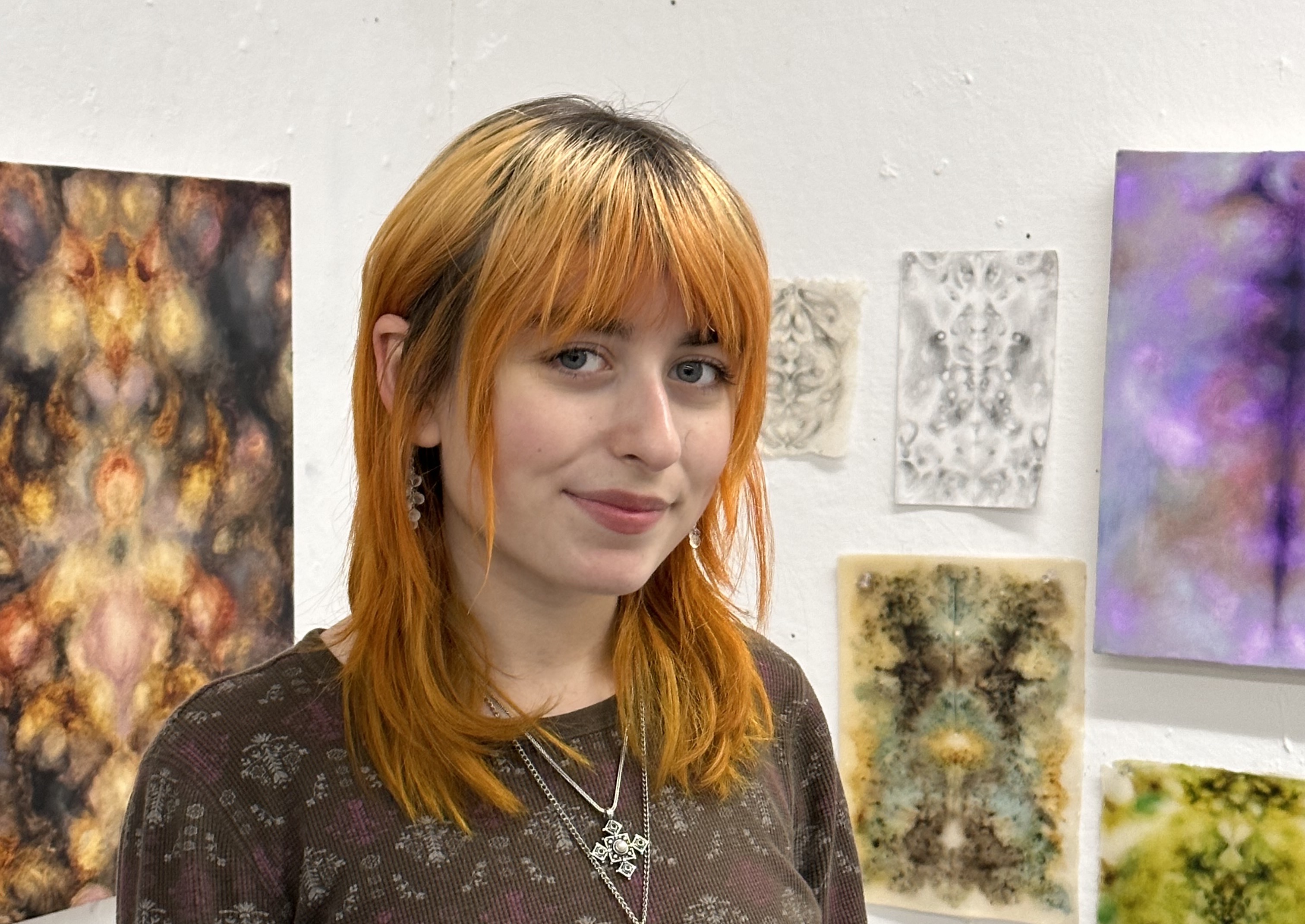 A person with bleached blonde and orange hair standing in front of painted canvasses hung on a wall