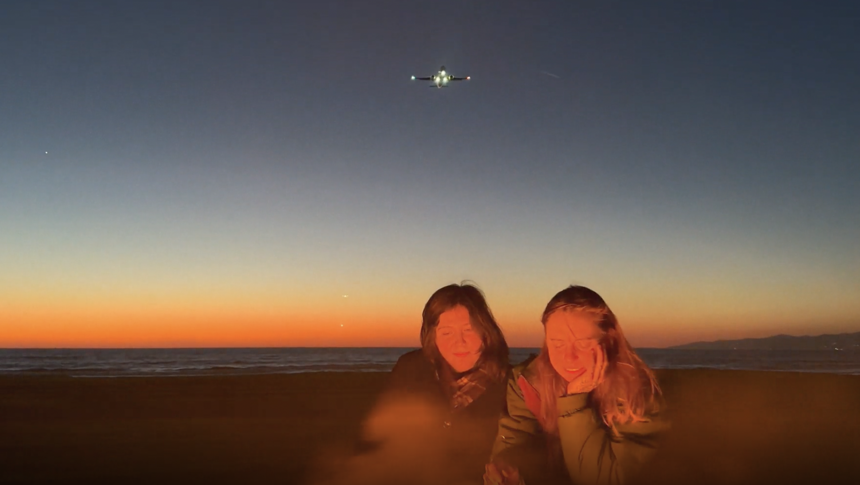 lucy dacus and julien baker on a beach at sunset as a plane flies overhead