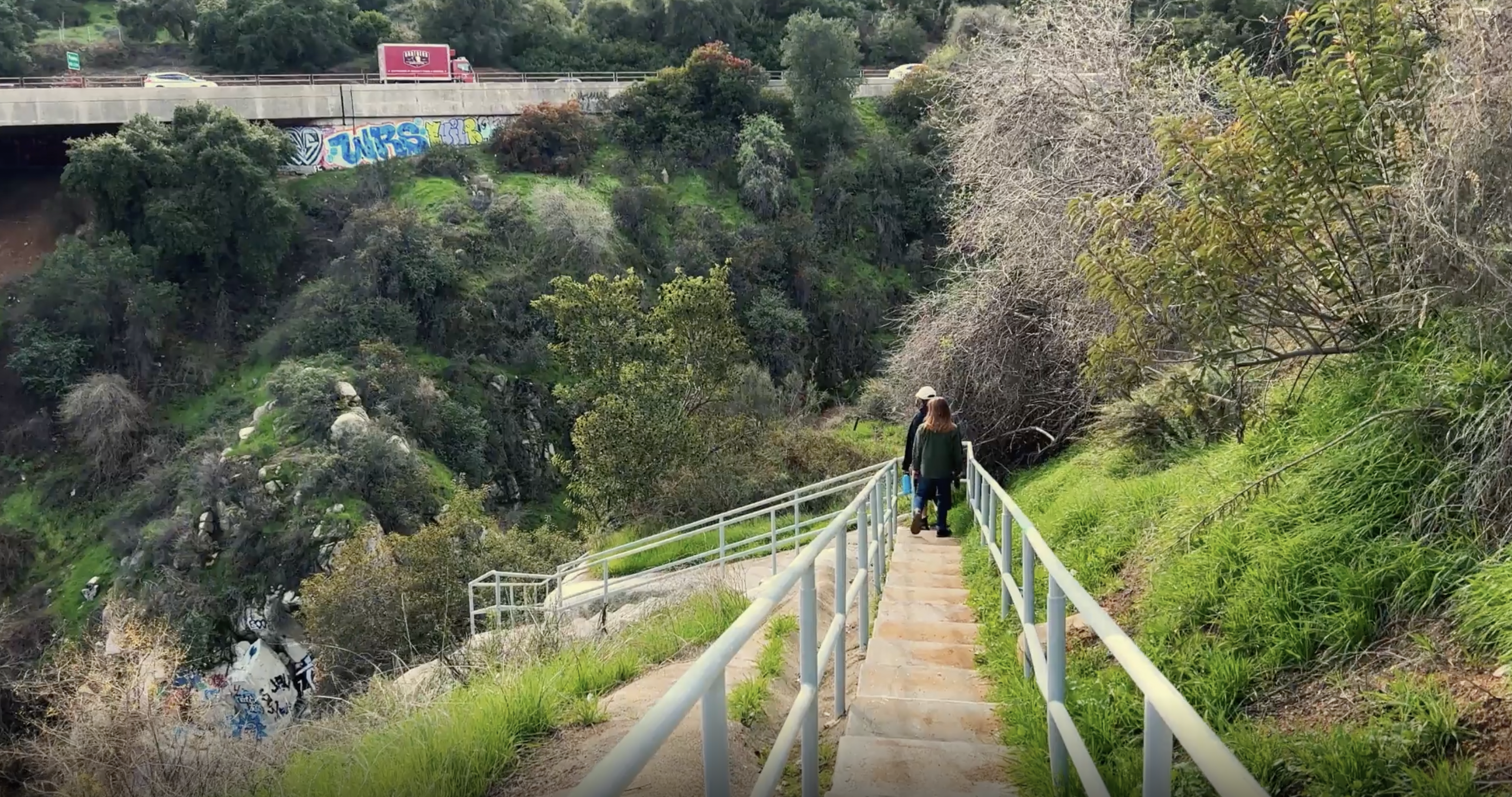 two people descend a staircase surrounded by greenery. a highway is in the background.