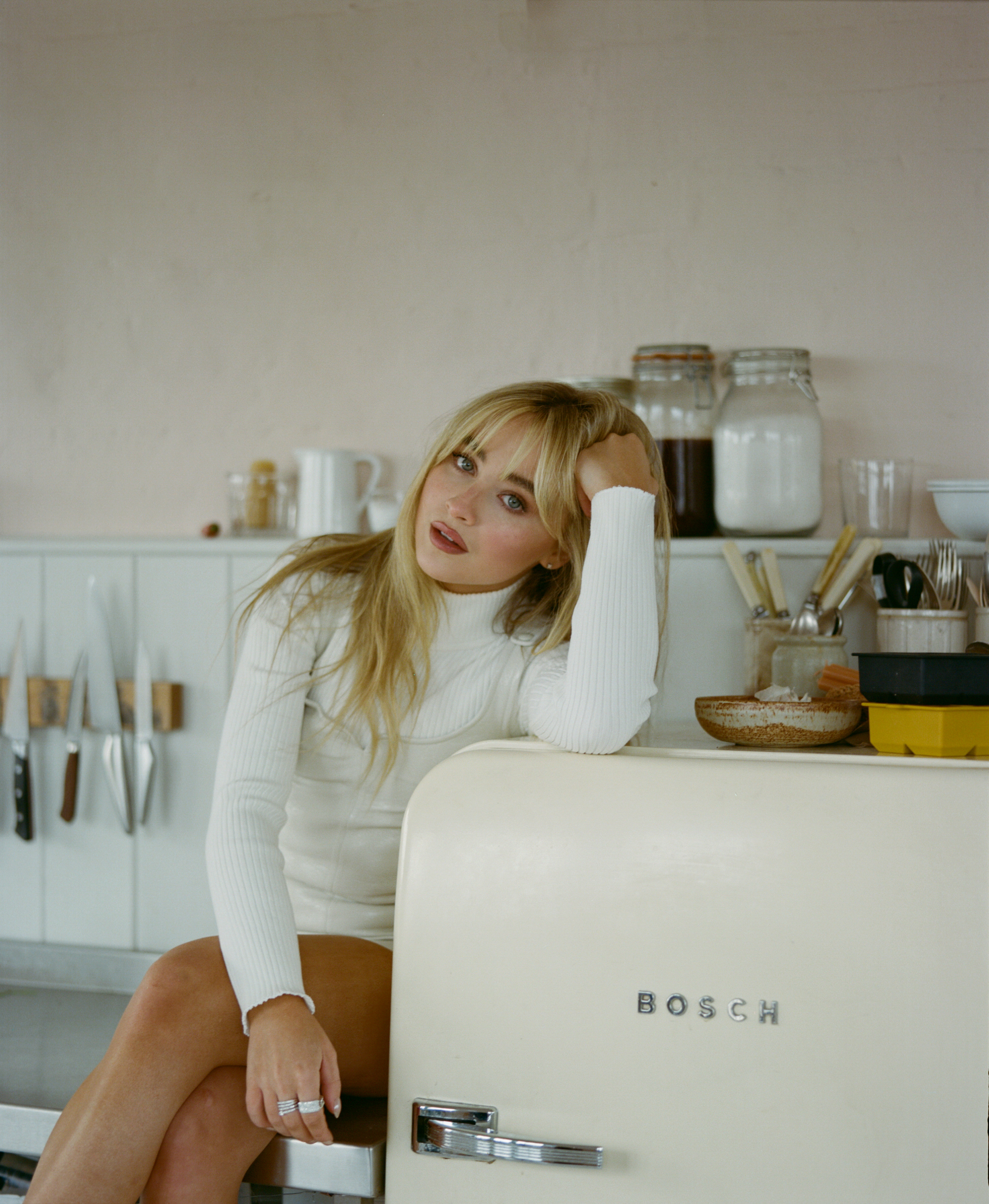sabrina carpenter, a blonde woman, sits on a kitchen countertop resting her arm on a fridge