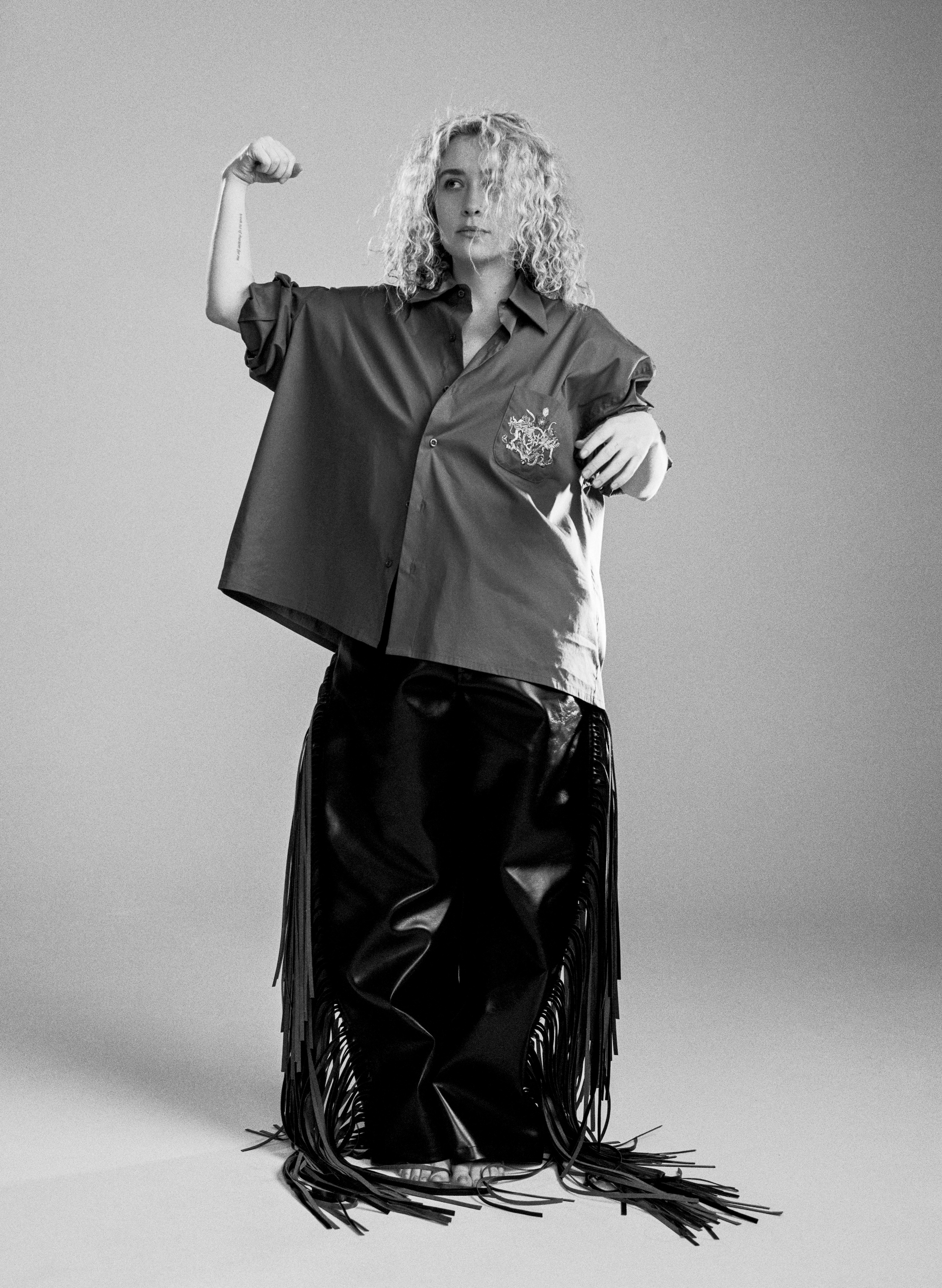 blondshell standing and making a fist wearing an oversized shirt and fringe trousers