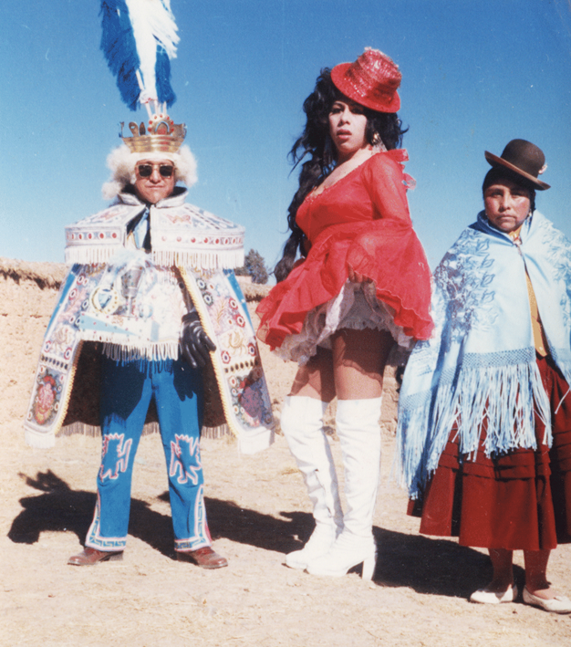 A travesti performer dressed as La China Morena against a desert backdrop, stood next to a man and woman in traditional Bolivian dress.