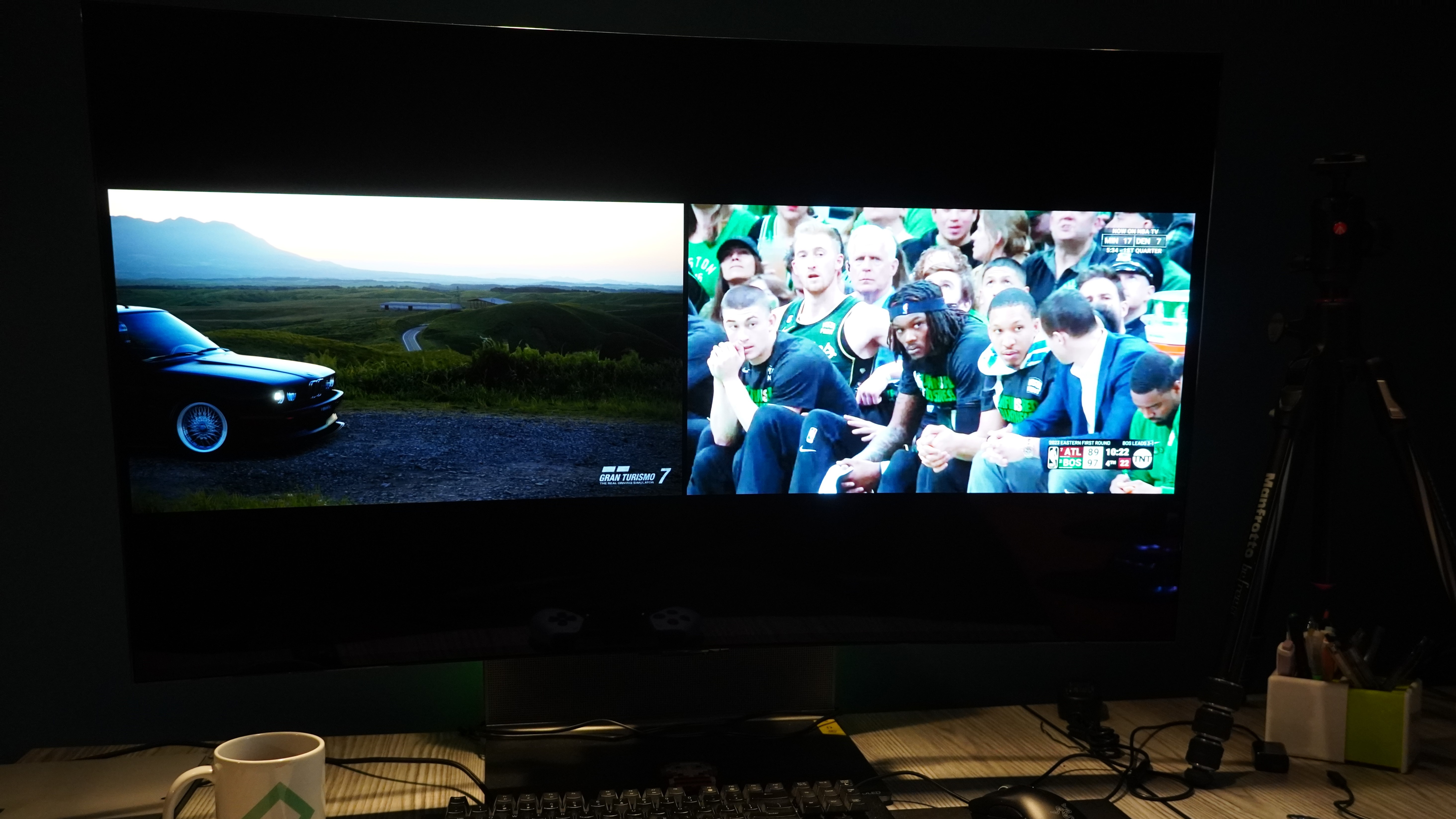 A split-screen mode on an OLED TV shows a BMW driving across a gravel road on the lefthand pane, which is showing Gran Turismo 7, while on the right hand pane a group of Boston Celtics players look gravely toward the camera during a close playoff game on YouTube TV.