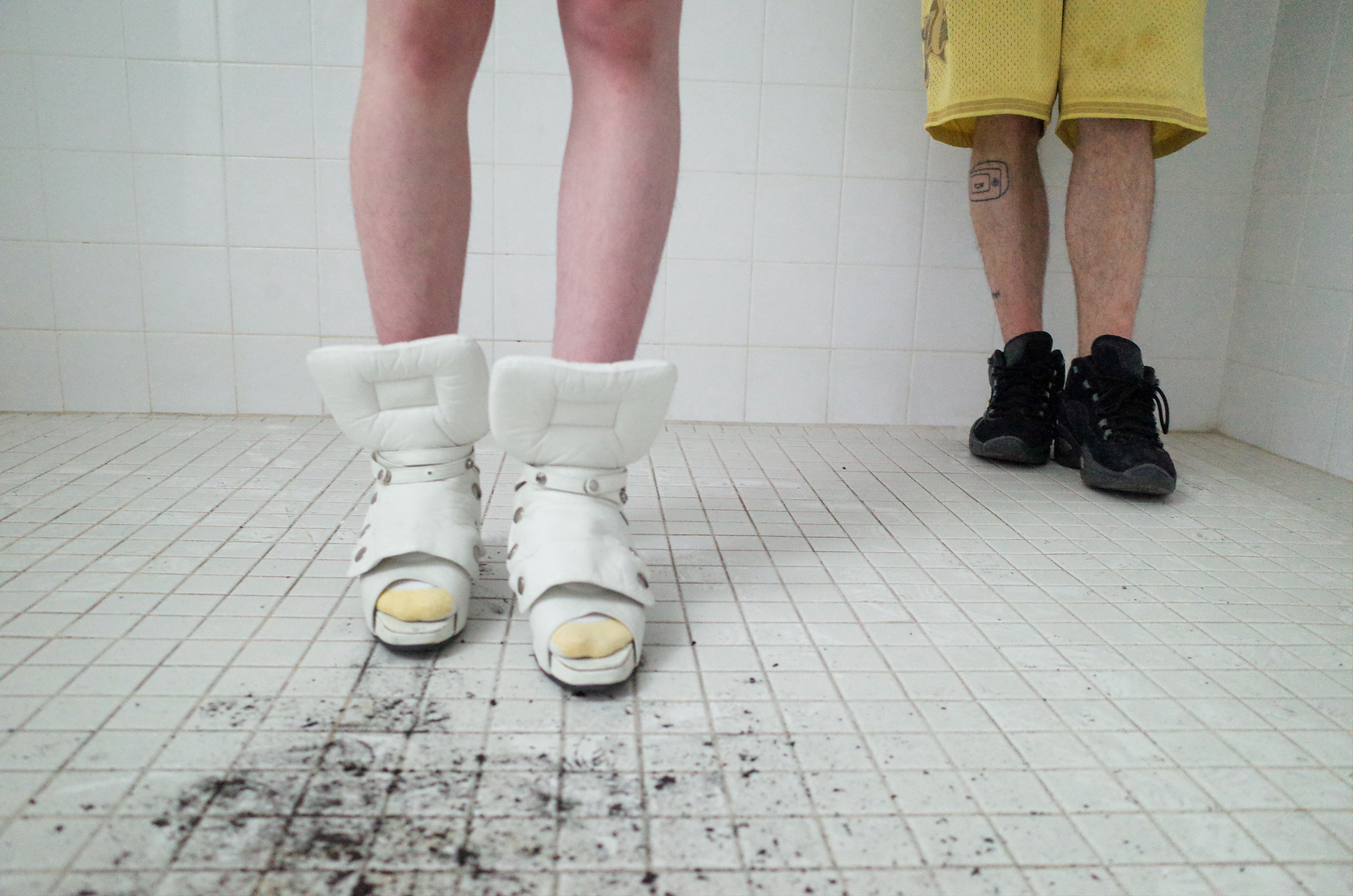 a close up of legs wearing oversized shoes in a white tiled shower room; there is mud on the floor