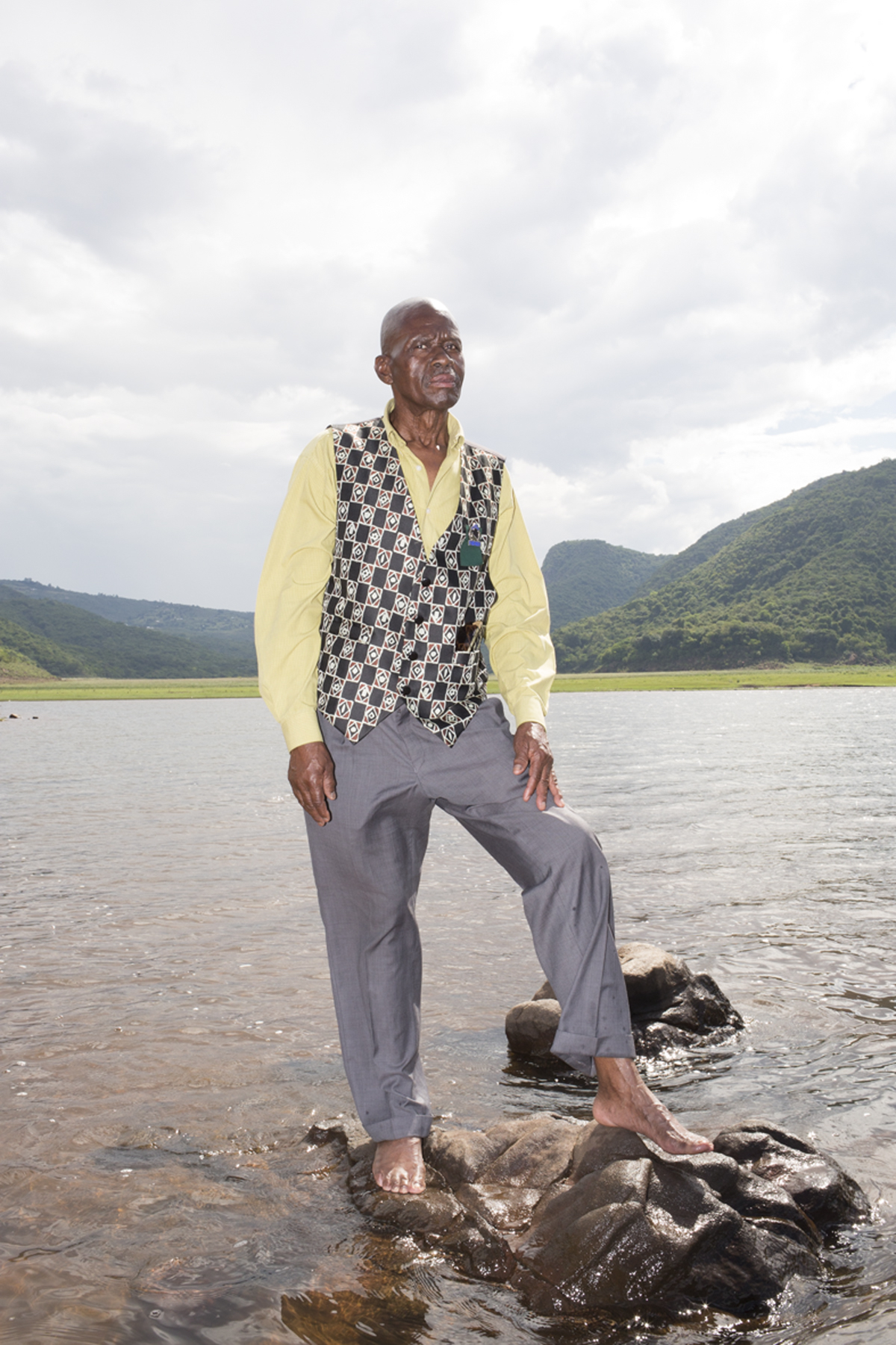 Giya Makondo-Wills, “They Came From The Water While The World Watched” – Elderly man wearing a yellow shirt, a chequered waistcoat and tailored grey trousers, standing barefoot on a rock in the middle of a river