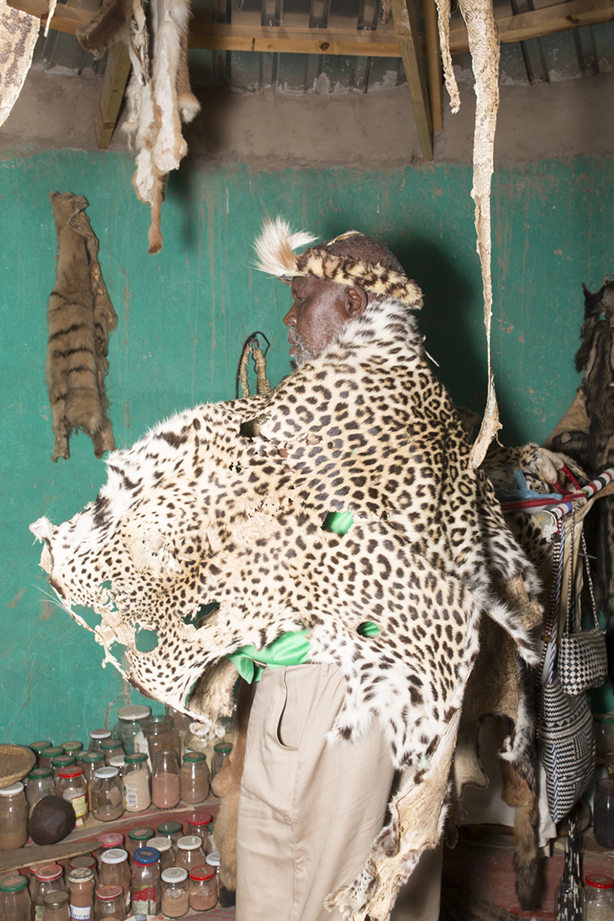 Giya Makondo-Wills, “They Came From The Water While The World Watched” – Profile of a man wearing a leopard skin and headband, inside a hut with lots of spices and animal skins hanging from the walls and the ceiling