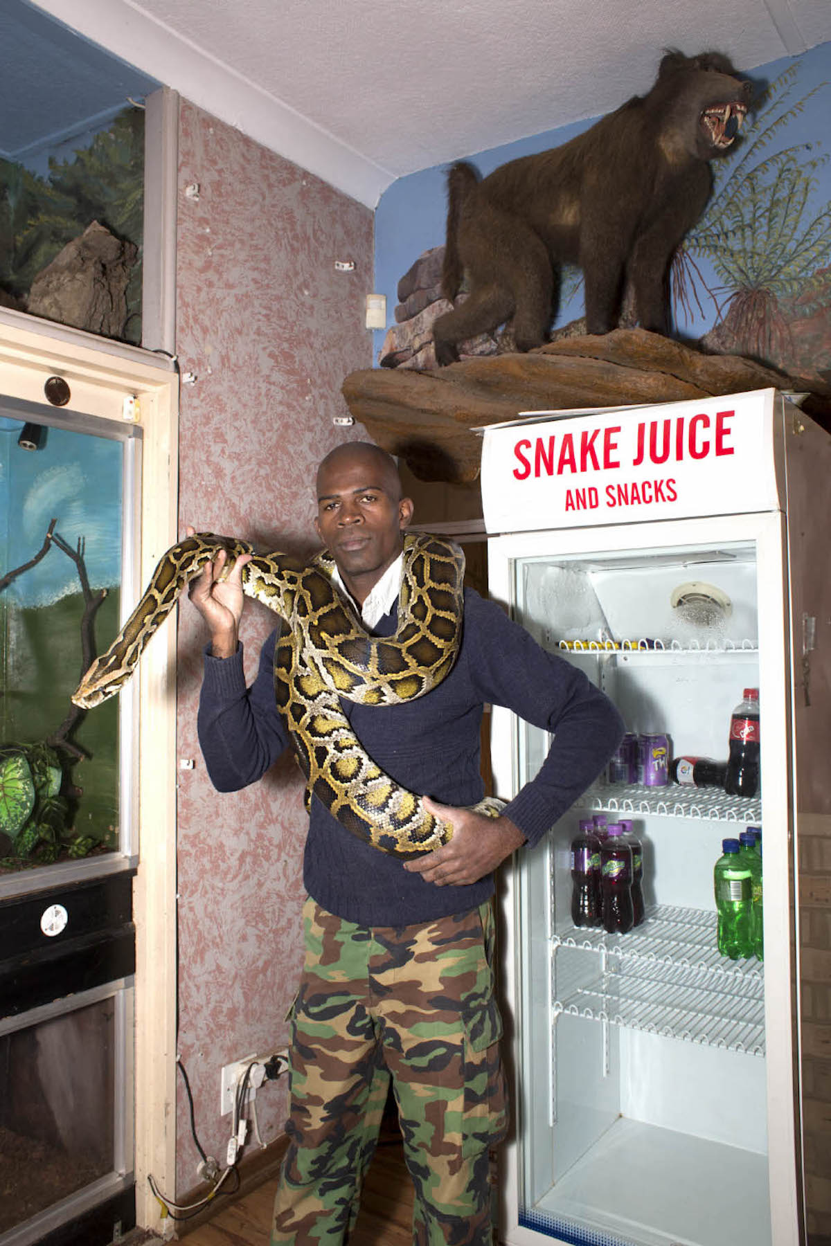 Giya Makondo-Wills, “They Came From The Water While The World Watched” – bald man wearing a blue jumper and military pants with a large snake wrapped around their neck and torso, standing in front of a refrigerator with a sign reading 
