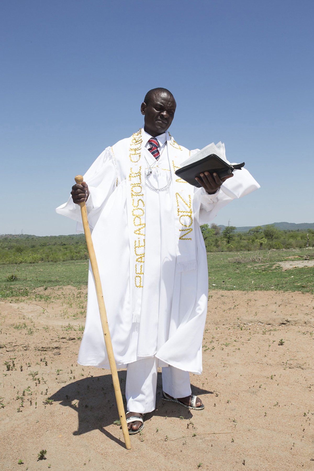 Giya Makondo-Wills, “They Came From The Water While The World Watched” – Pastor dressed in white and gold, holding a cane and a bible in a field