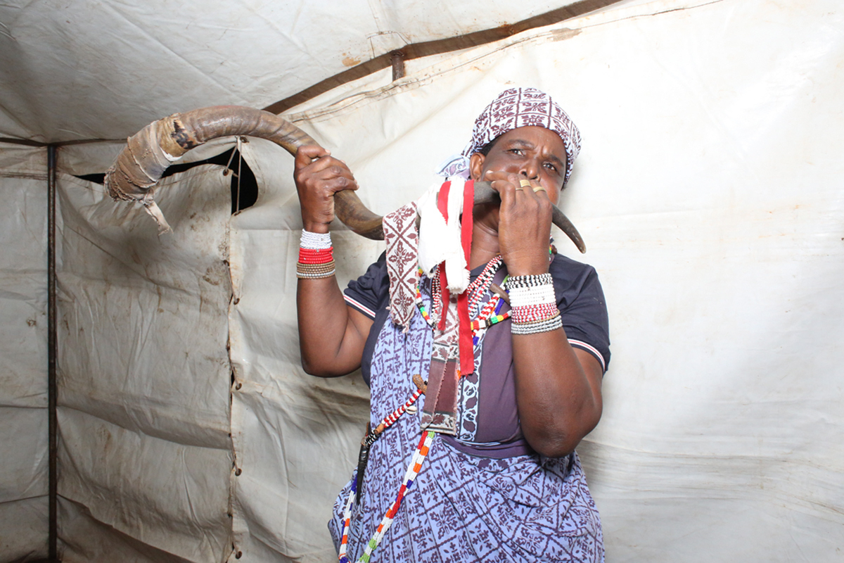 Giya Makondo-Wills, “They Came From The Water While The World Watched” – middle-aged woman inside a tent, blowing on a large animal horn
