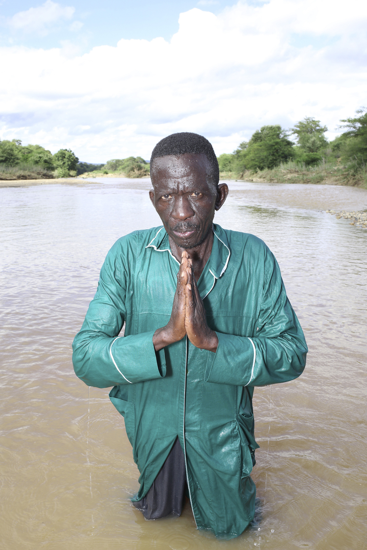 Giya Makondo-Wills, “They Came From The Water While The World Watched” – middle-aged man with a wet long green robe standing in a river and holding his hands in prayer